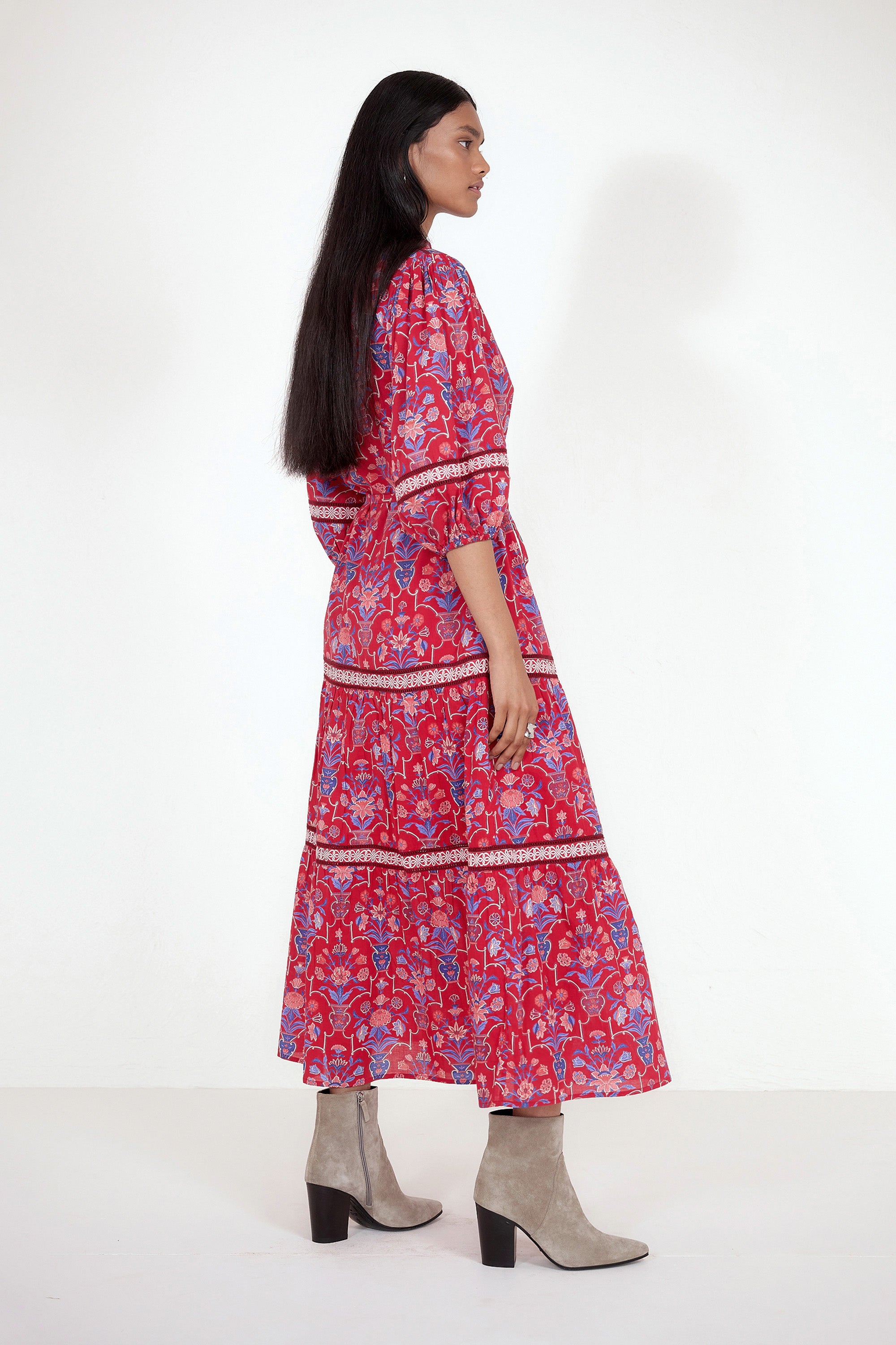 Side view of model standing. She is wearing the Liza dress in Floral Vase print. It features a tie neckline, soft ruffles, lace sleeve and skirt details, and a fabric belt.