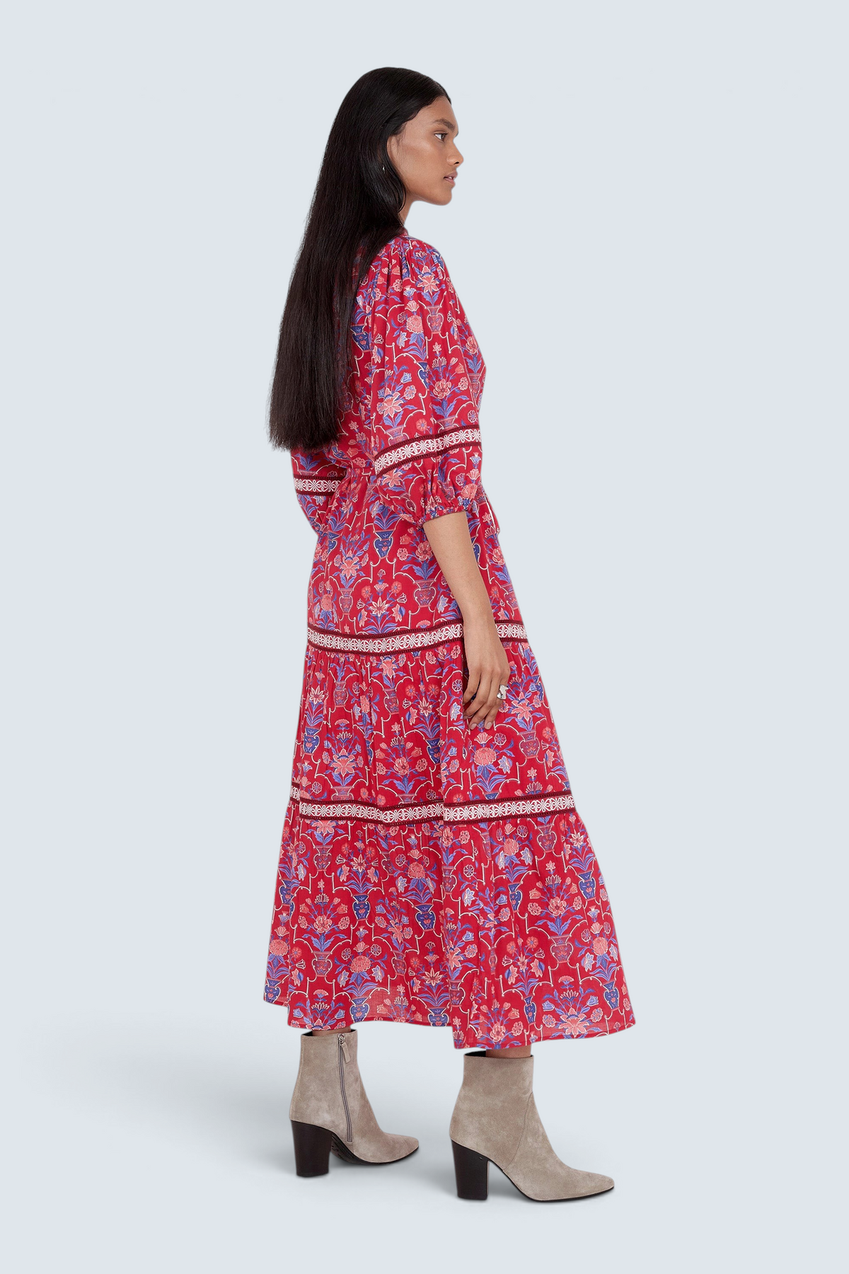 Side view of model standing. She is wearing the Liza dress in Floral Vase print. It features a tie neckline, soft ruffles, lace sleeve and skirt details, and a fabric belt.