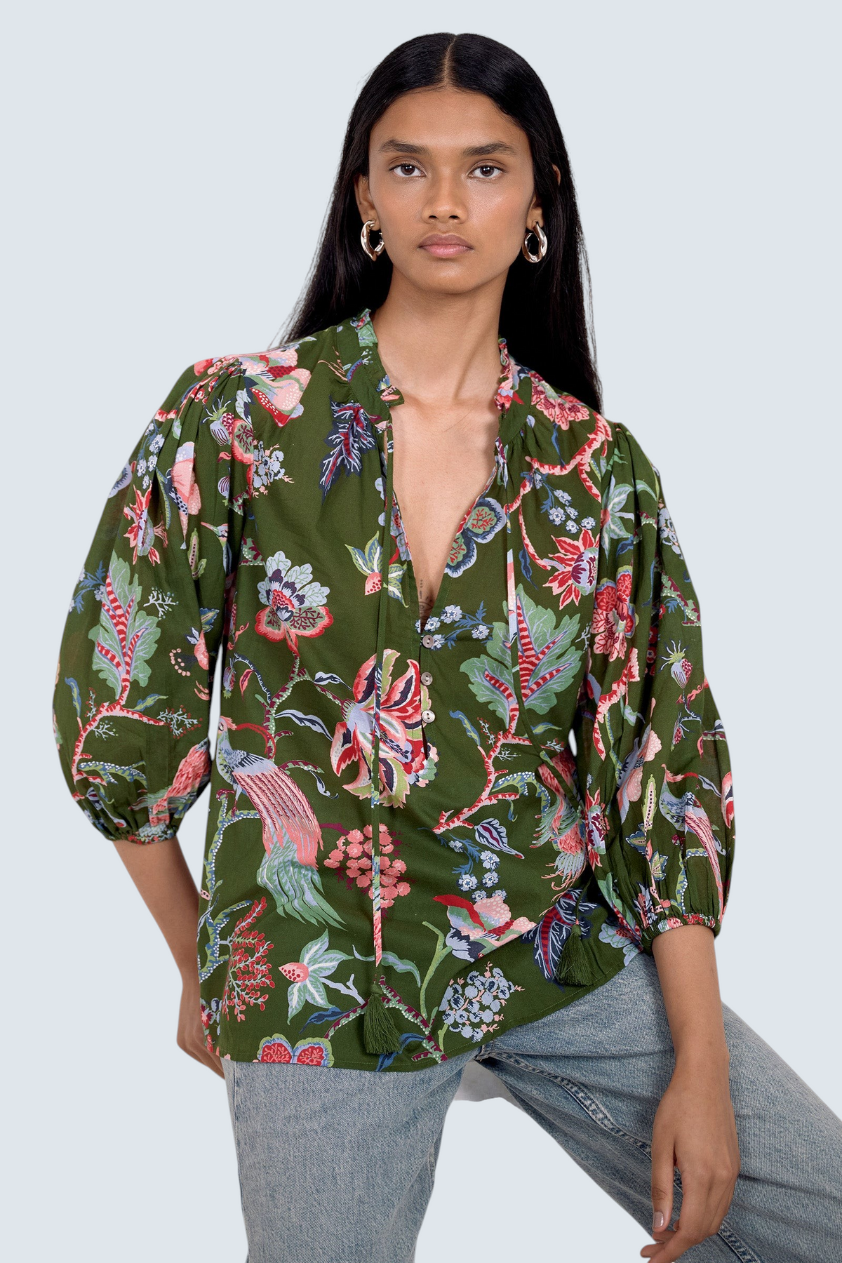 Front view of model sitting on chair. She is  wearing the Lorna top in Chintz print, featuring a V-neck with a soft ruffle and tie, a loose flowing body, and slightly voluminous sleeves, paired with jeans.