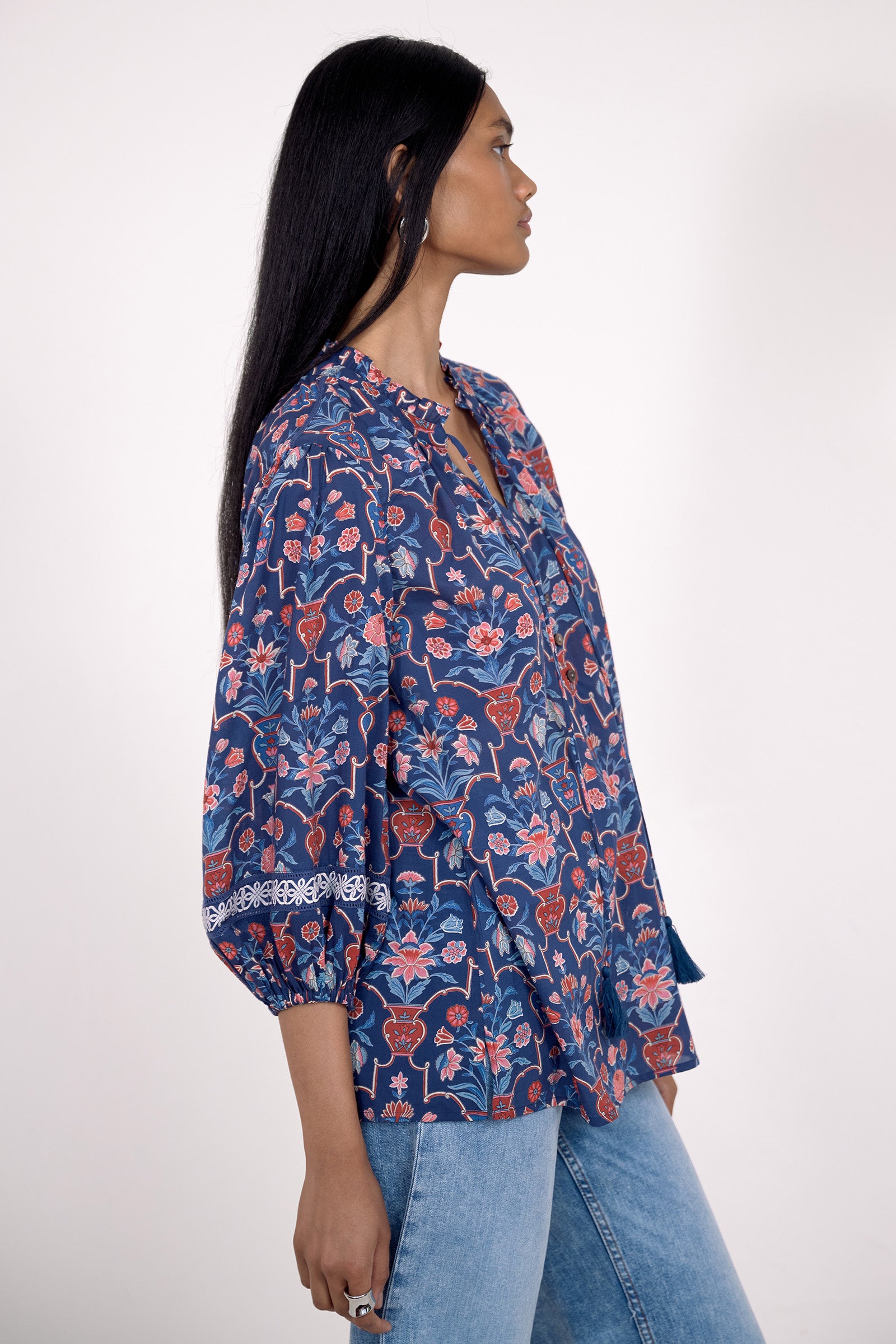 Side view of model standing. She is  wearing the Lorna top in Floral Vase print, featuring a V-neck with a soft ruffle and tie, lace trim inset at the sleeves, a loose flowing body, and slightly voluminous sleeves, paired with jeans.