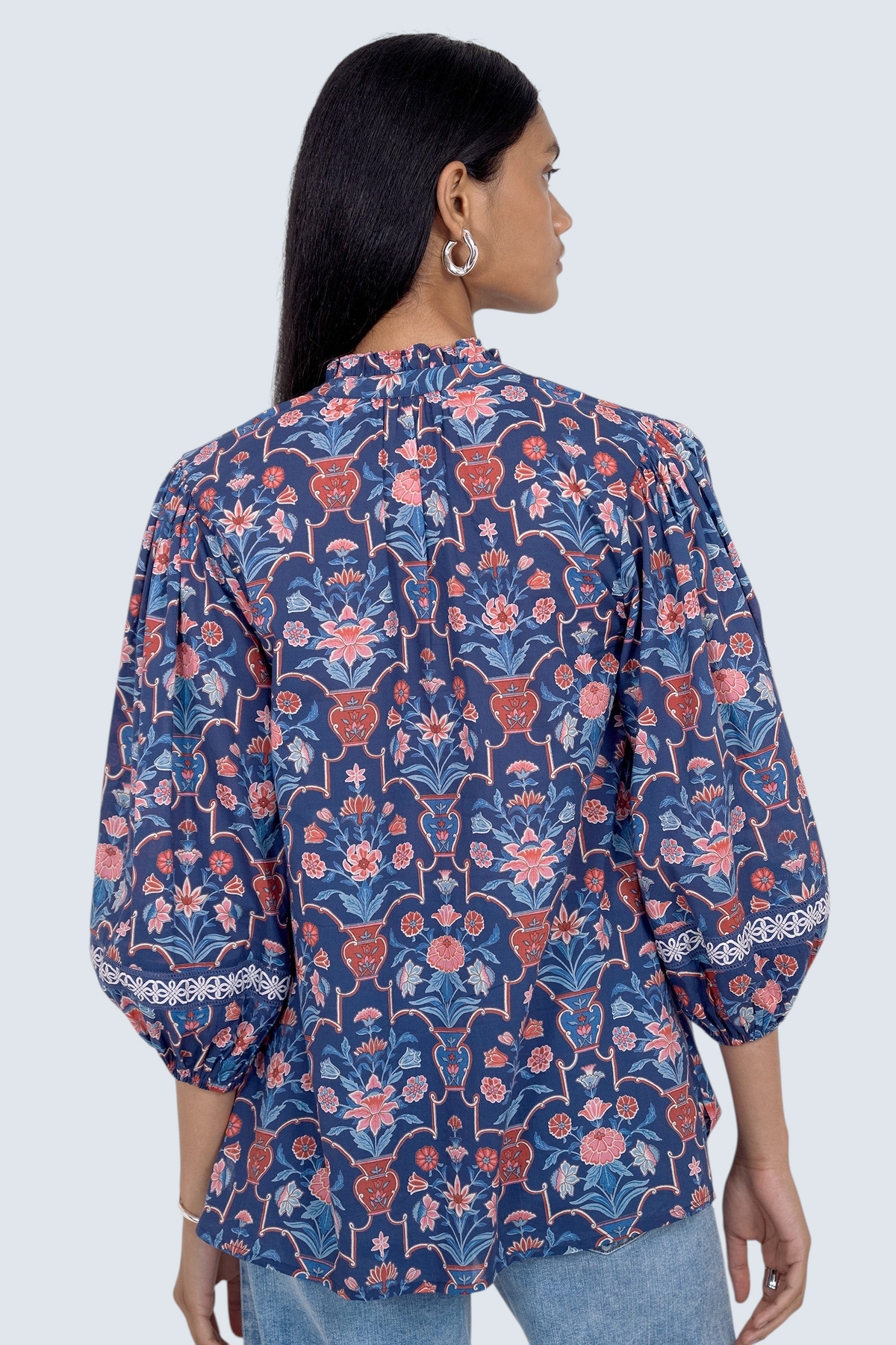 Back view of model standing. She is  wearing the Lorna top in Floral Vase print, featuring a V-neck with a soft ruffle and tie, lace trim inset at the sleeves, a loose flowing body, and slightly voluminous sleeves, paired with jeans.