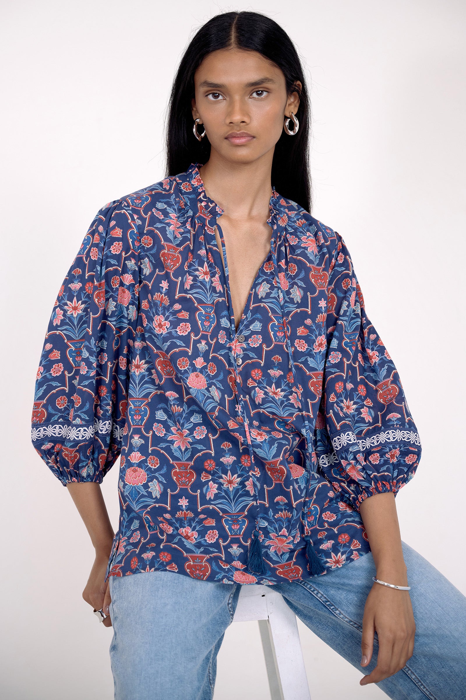 Front view of model sitting on a chair. She is wearing the Lorna top in Floral Vase print, featuring a V-neck with a soft ruffle and tie, lace trim inset at the sleeves, a loose flowing body, and slightly voluminous sleeves, paired with jeans.