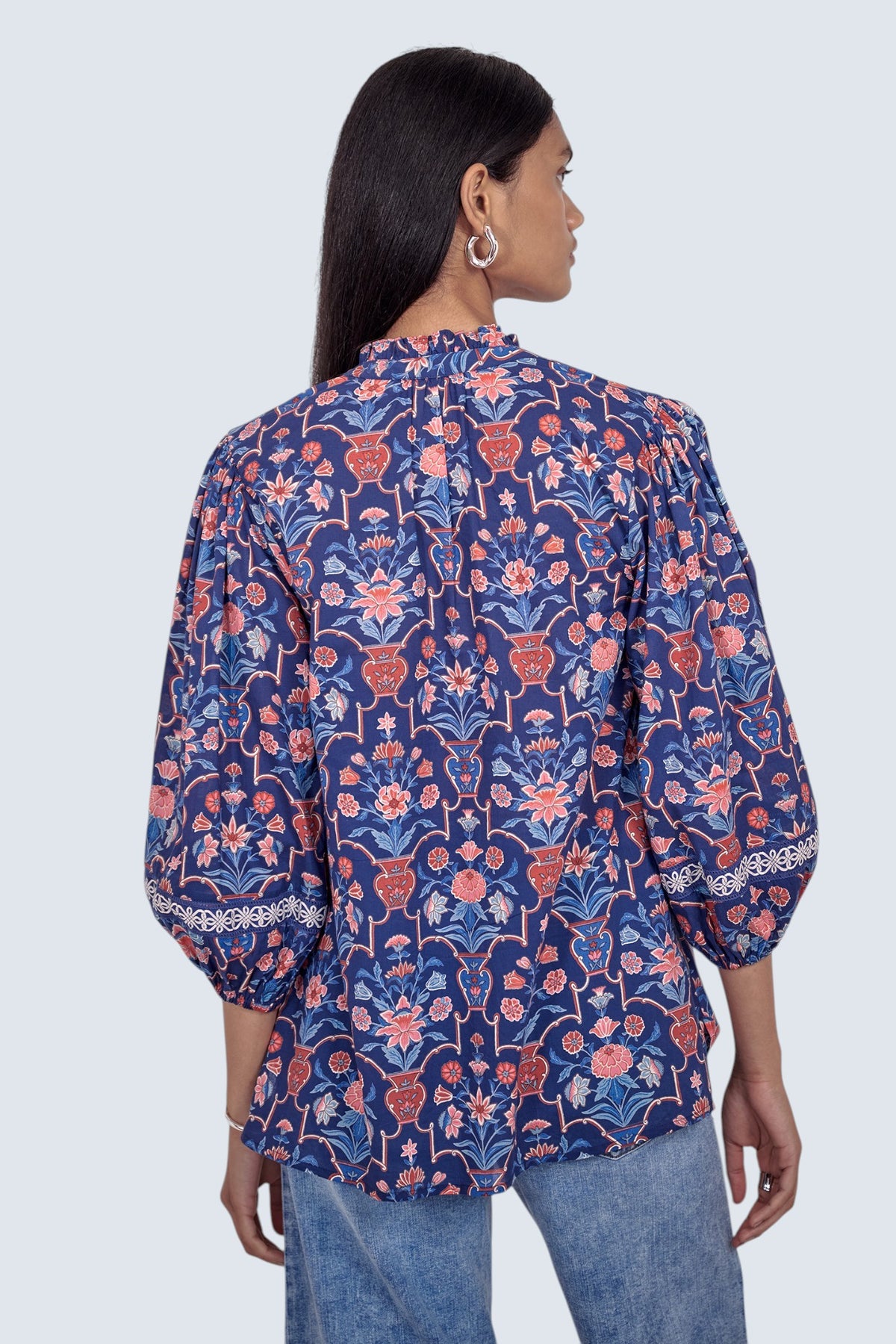 Back view of model standing wearing the Lorna top in Floral Vase print, featuring a V-neck with ruffle and tie, lace-trimmed sleeves, and a loose flowing silhouette, paired with jeans.