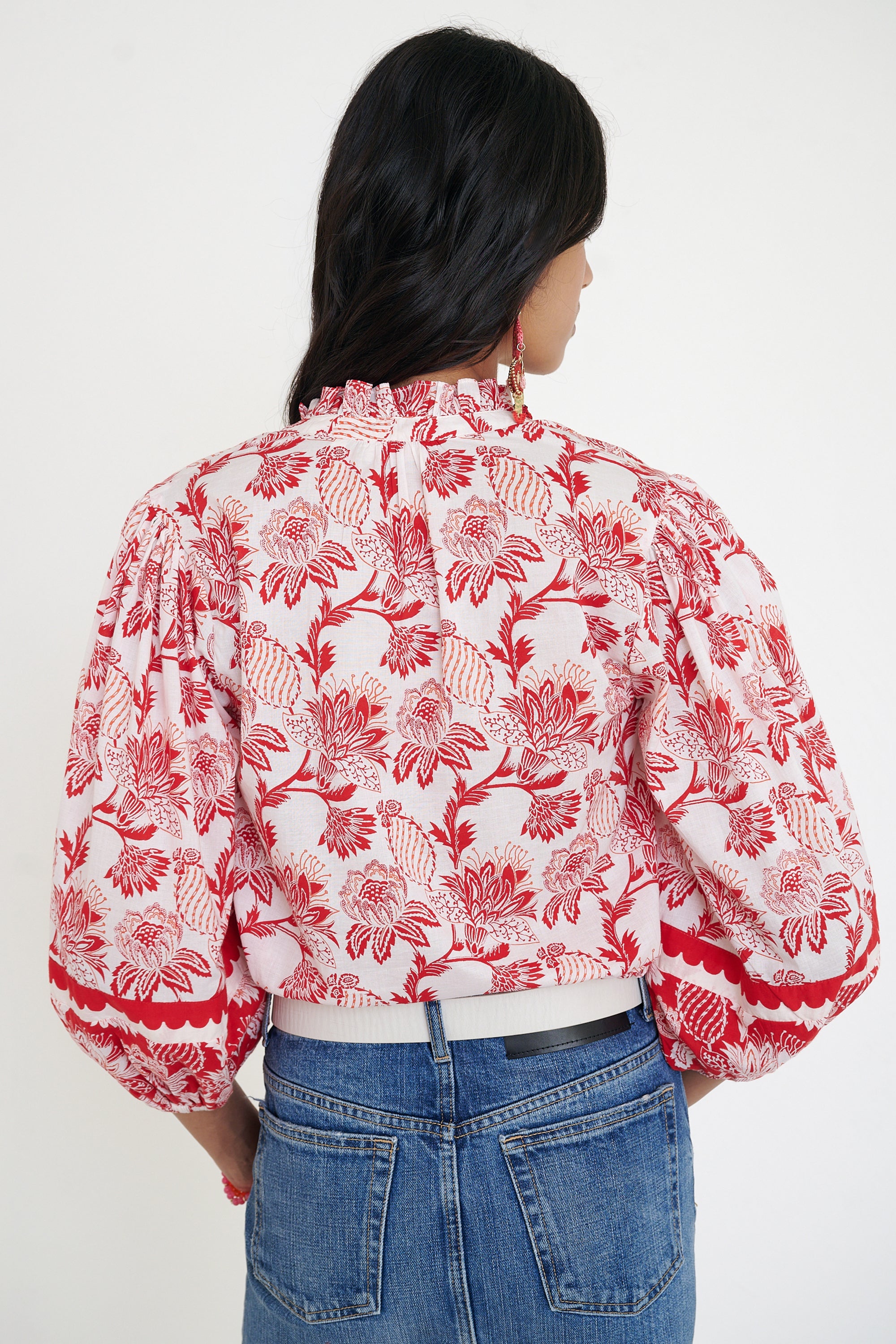 Back view of woman standing. She is  wearing the Lorna top in the Lizzie print, featuring a V-neck with a soft ruffle, adjustable tie closure, voluminous sleeves, and winding floral vine motifs.