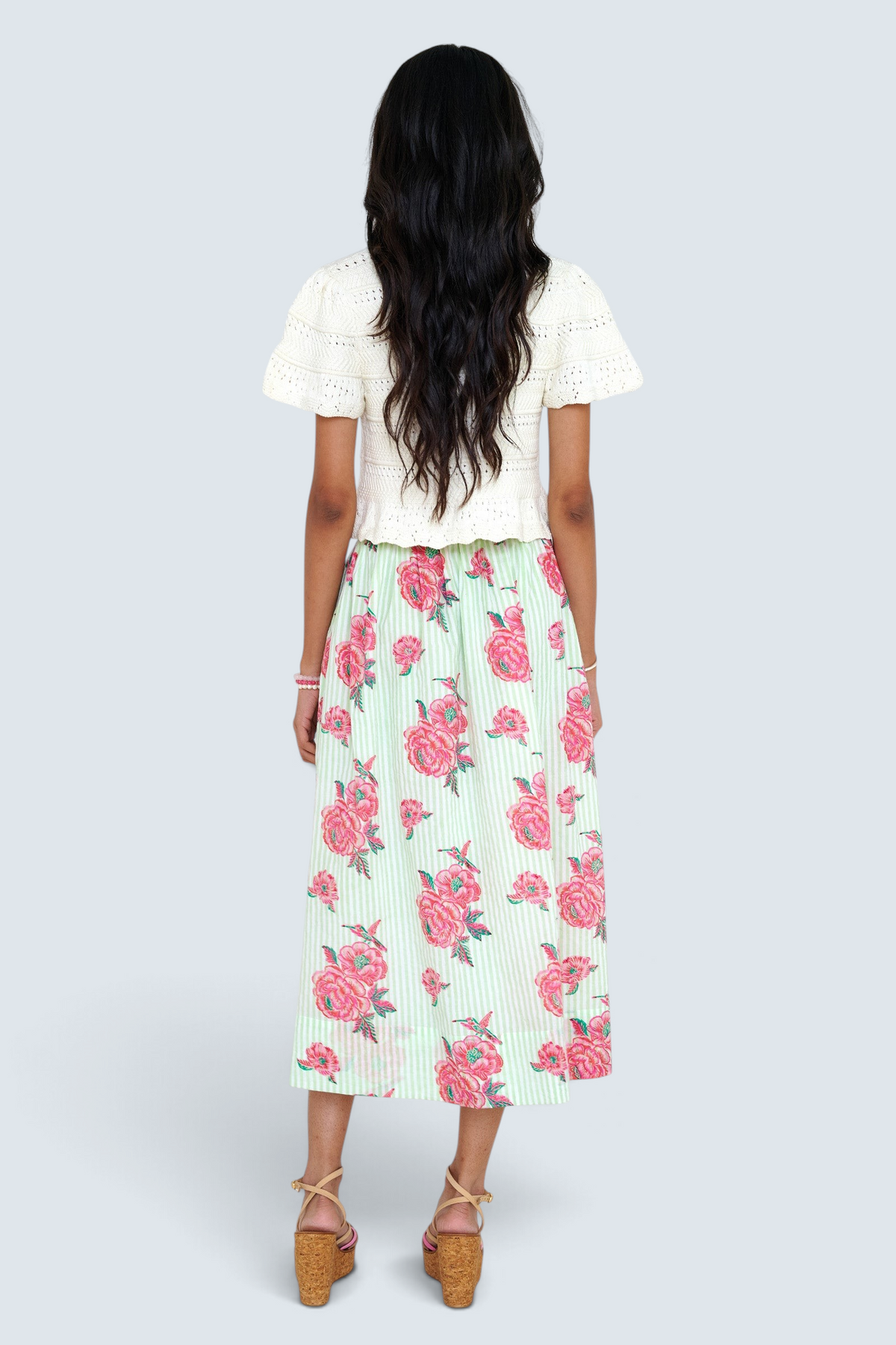 Back view of a woman standing wearing the Magdalena skirt in Eliza Rose print. It features all-around pleats, side pockets, and a side zipper closure with hand block-printed detailing. She paired it with our Laura crochet cardigan.
