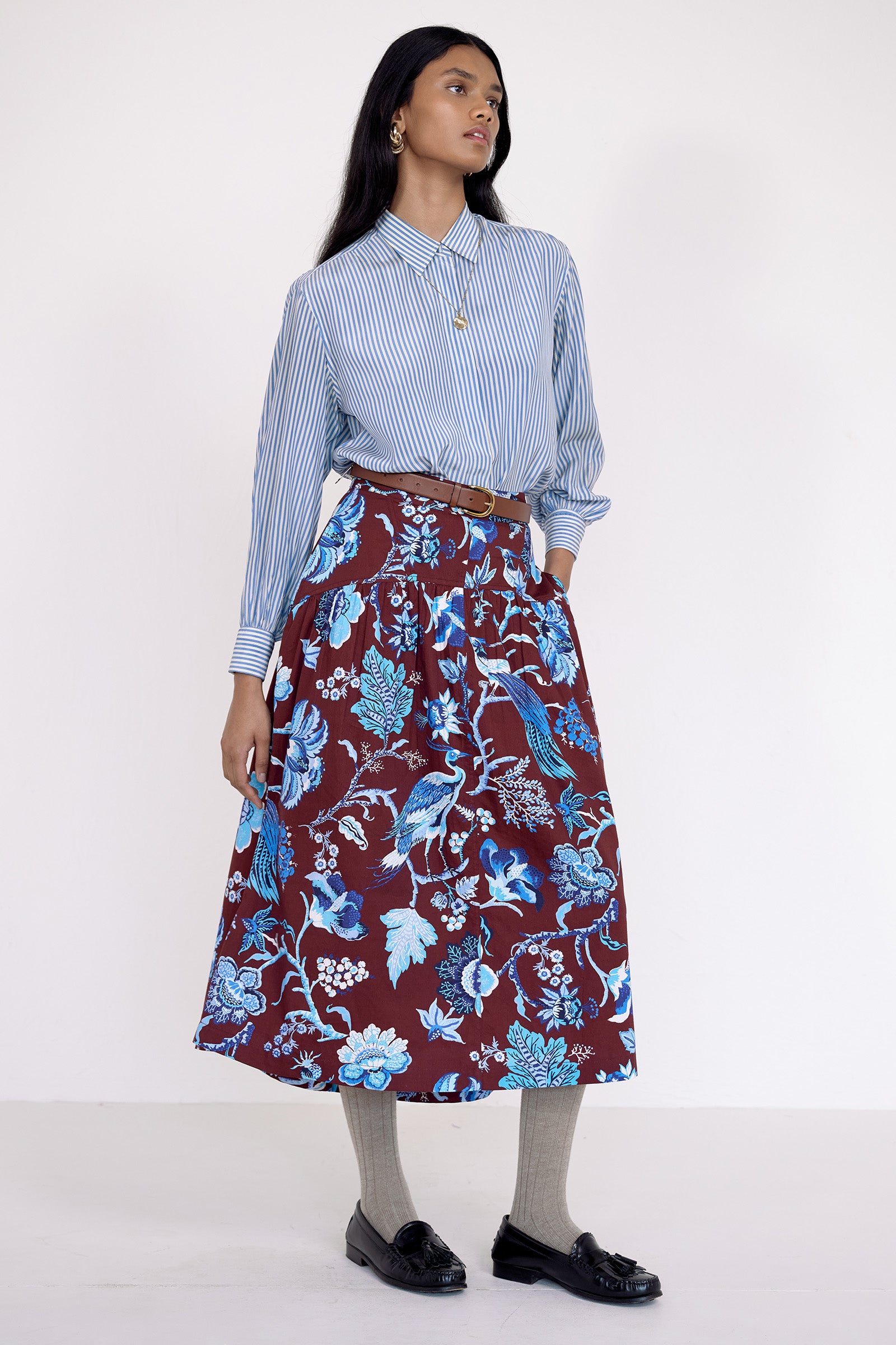 Front side view of model standing. She wearing the Melodie skirt in Chintz print, featuring a slim waist, gathered silhouette, and side-seam pockets.Its paired with a pinstripe shirt, belt, socks, shoe and accessories.