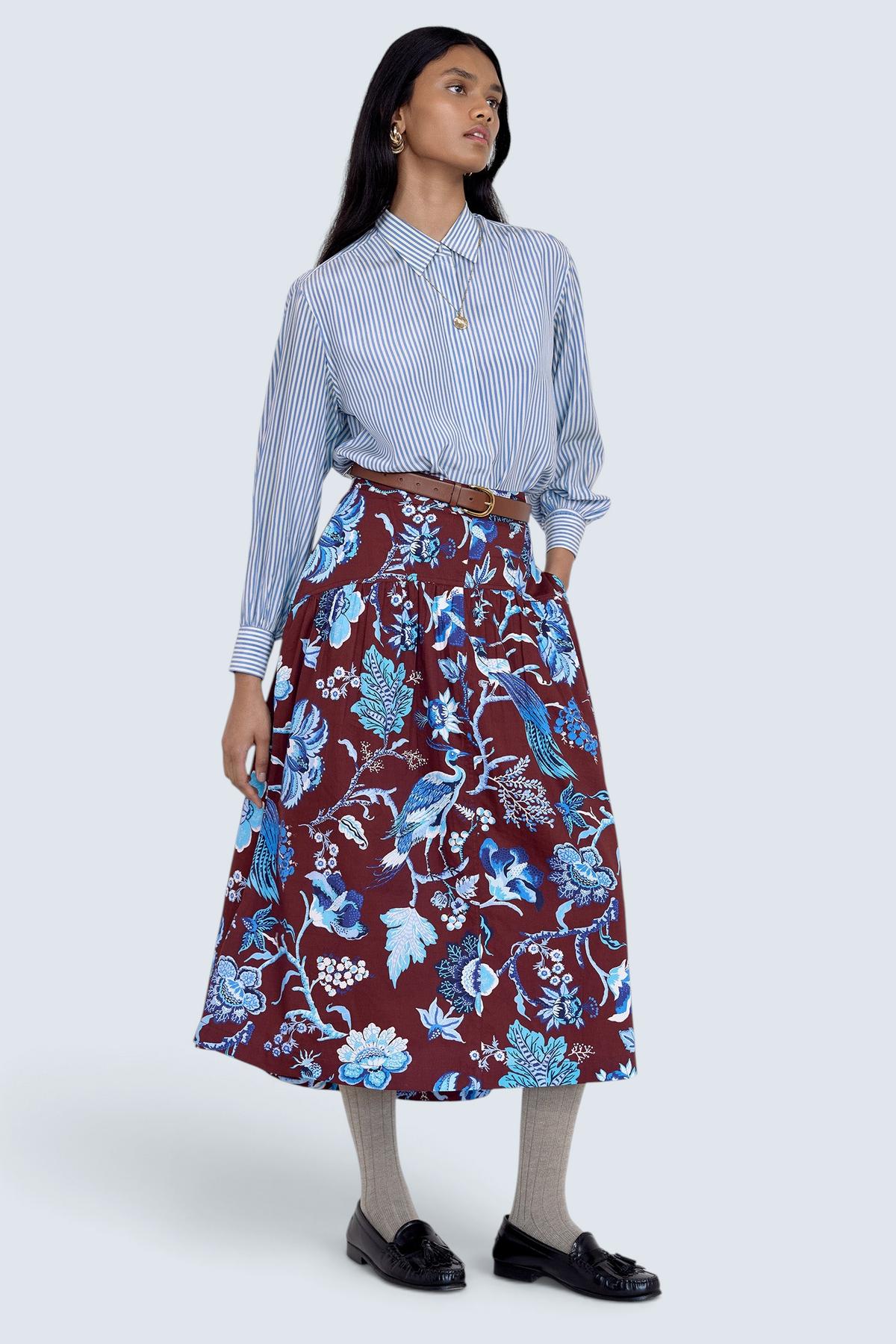 Front side view of model standing. She wearing the Melodie skirt in Chintz print, featuring a slim waist, gathered silhouette, and side-seam pockets.Its paired with a pinstripe shirt, belt, socks, shoe and accessories.