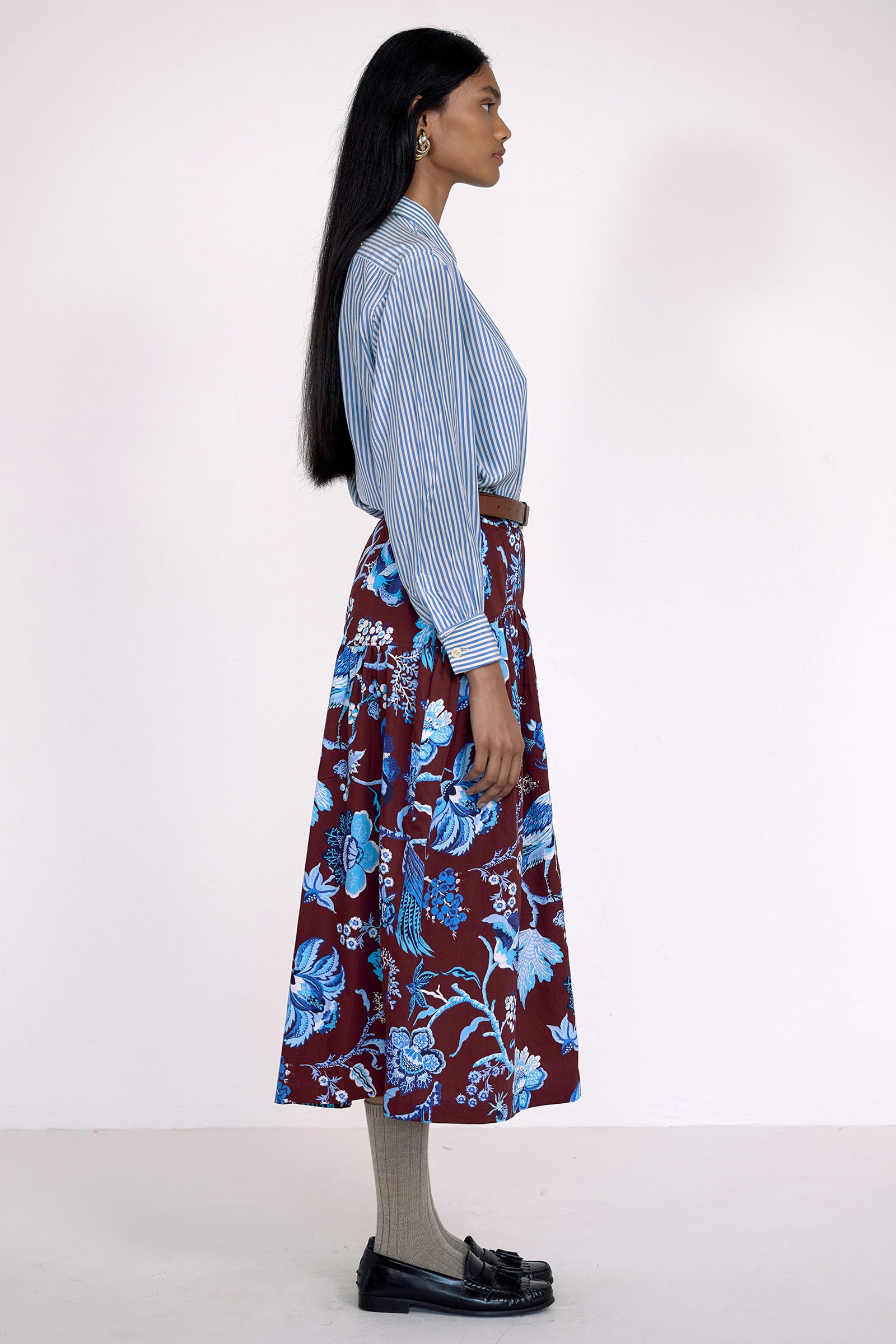 Side view of model standing. She wearing the Melodie skirt in Chintz print, featuring a slim waist, gathered silhouette, and side-seam pockets.Its paired with a pinstripe shirt, belt, socks, shoe and accessories.