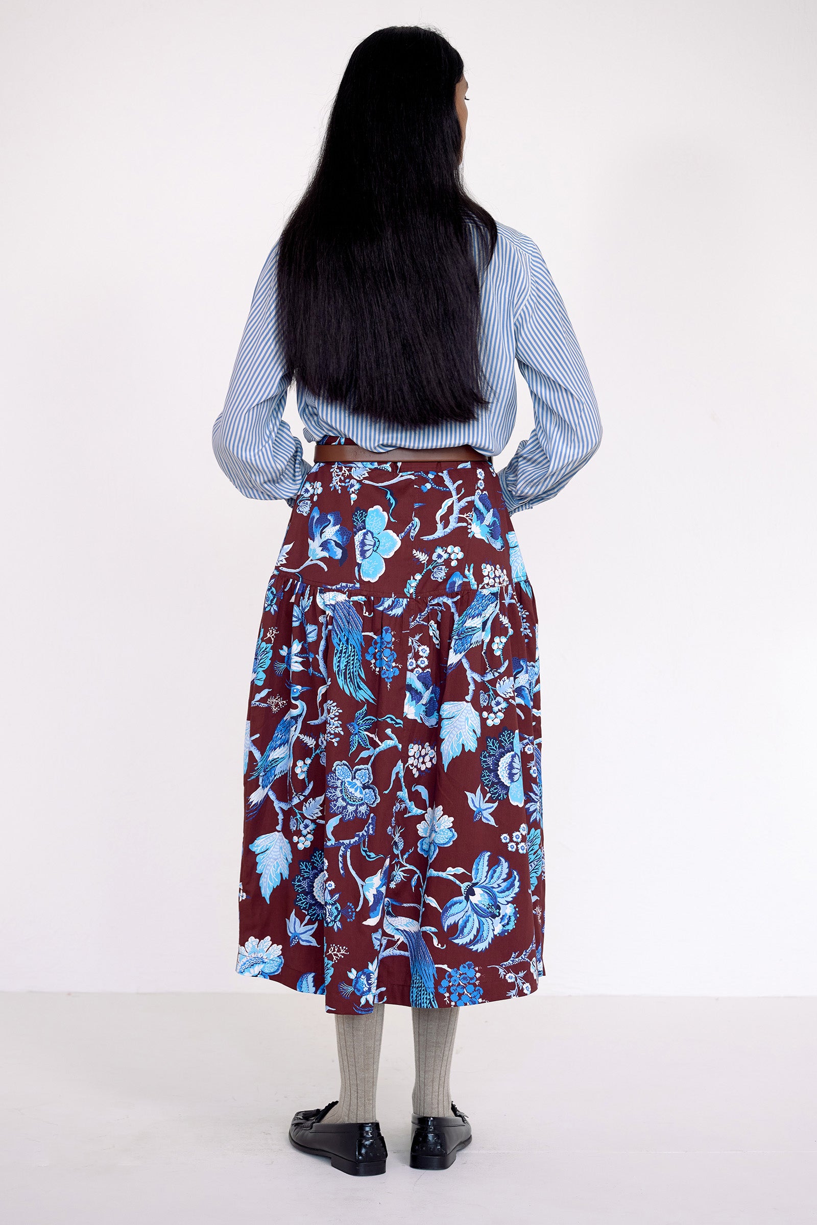 Back view of model standing. She wearing the Melodie skirt in Chintz print, featuring a slim waist, gathered silhouette, and side-seam pockets.Its paired with a pinstripe shirt, belt, socks, shoe and accessories.