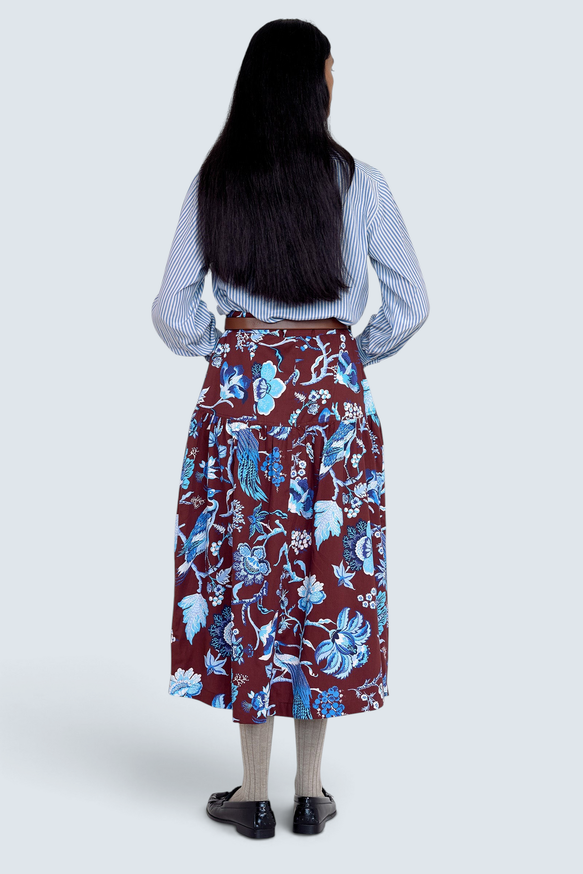 Back view of model standing. She wearing the Melodie skirt in Chintz print, featuring a slim waist, gathered silhouette, and side-seam pockets.Its paired with a pinstripe shirt, belt, socks, shoe and accessories.