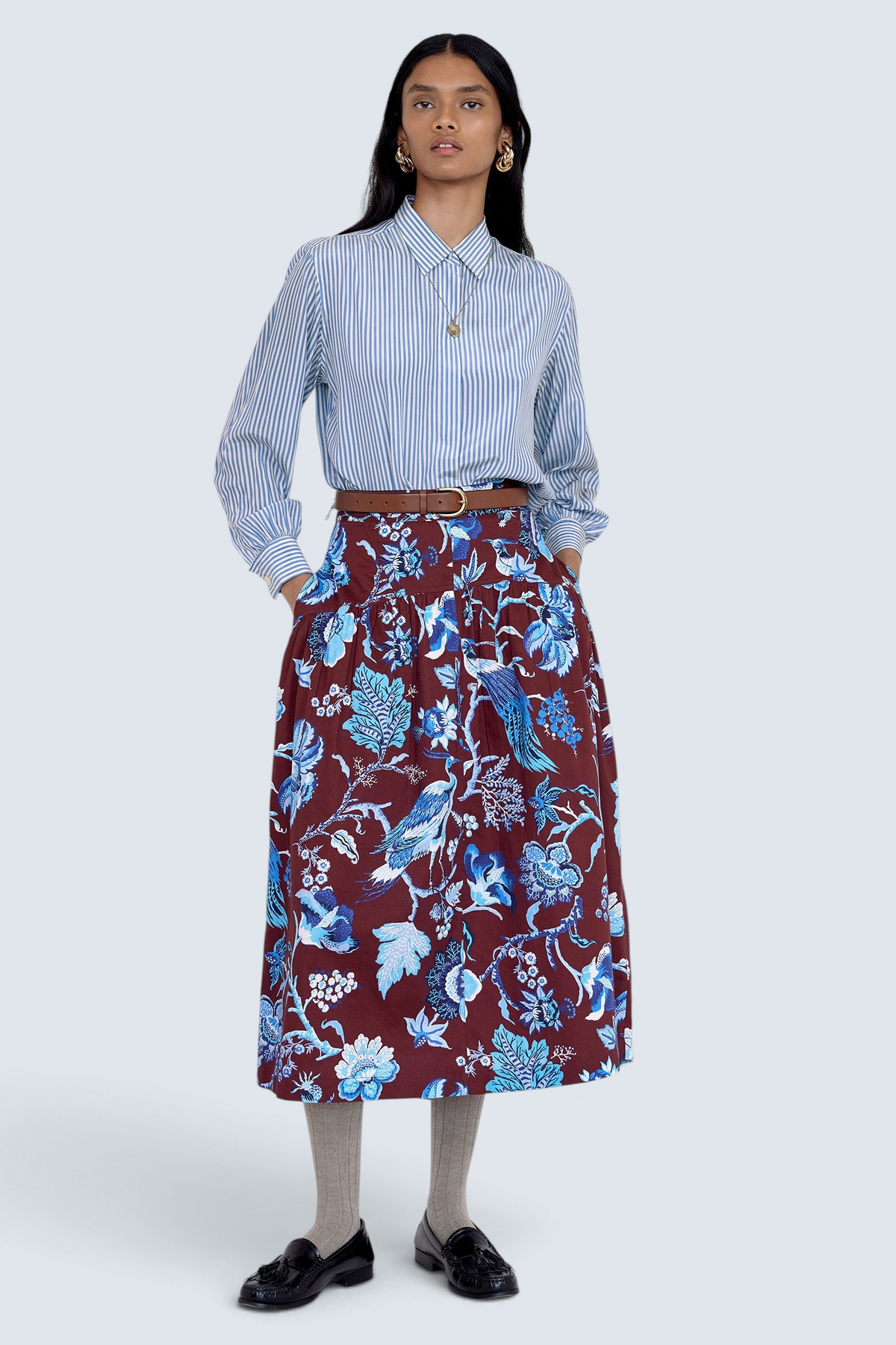 Front view of model standing. She wearing the Melodie skirt in Chintz print, featuring a slim waist, gathered silhouette, and side-seam pockets.Its paired with a pinstripe shirt, belt, socks, shoe and accessories.