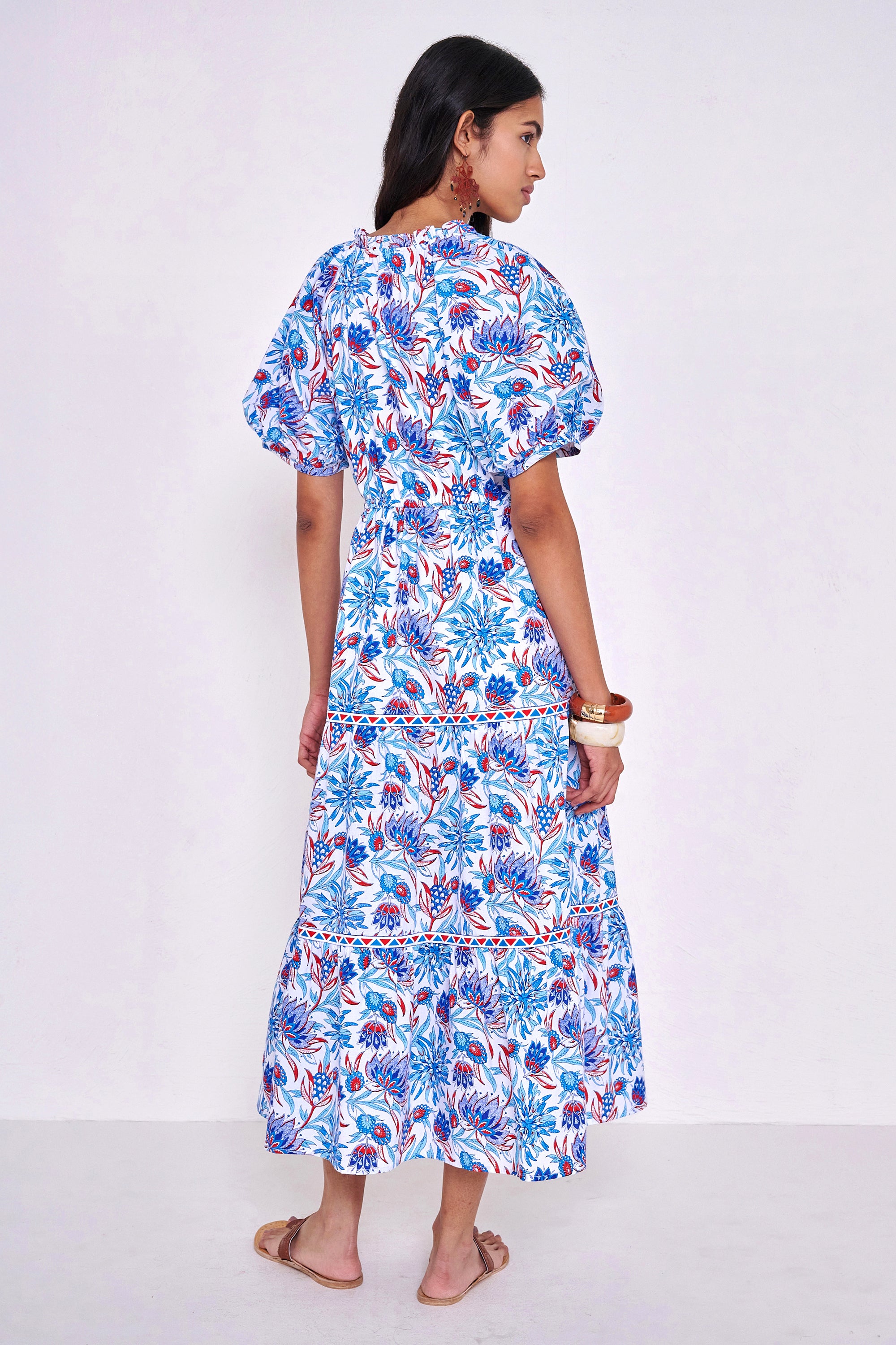 Back view of woman standing wearing the Poppy dress in the Philippa print, featuring raglan blouson sleeves, a button-front placket, a flowing skirt, and floral motifs inspired by the wildflowers of Provence.
