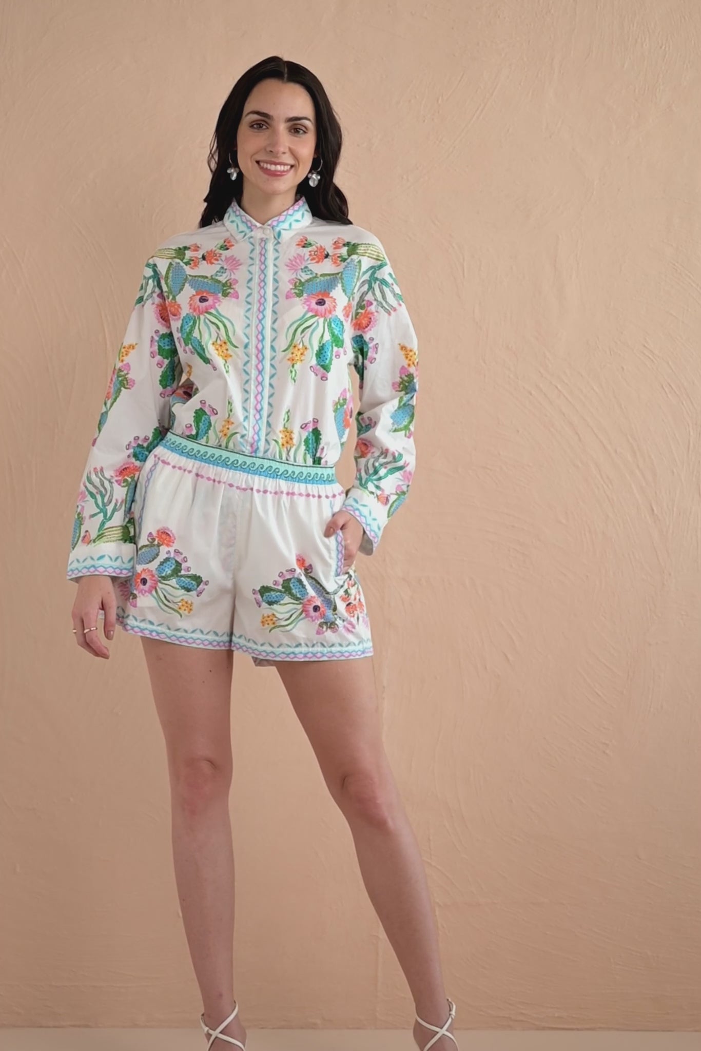 Video of a woman wearing a white co-ord set featuring a colorful Calypso Cacti Paradise print, including a long-sleeved button-down shirt and matching high-waisted shorts, styled with white strappy heels.