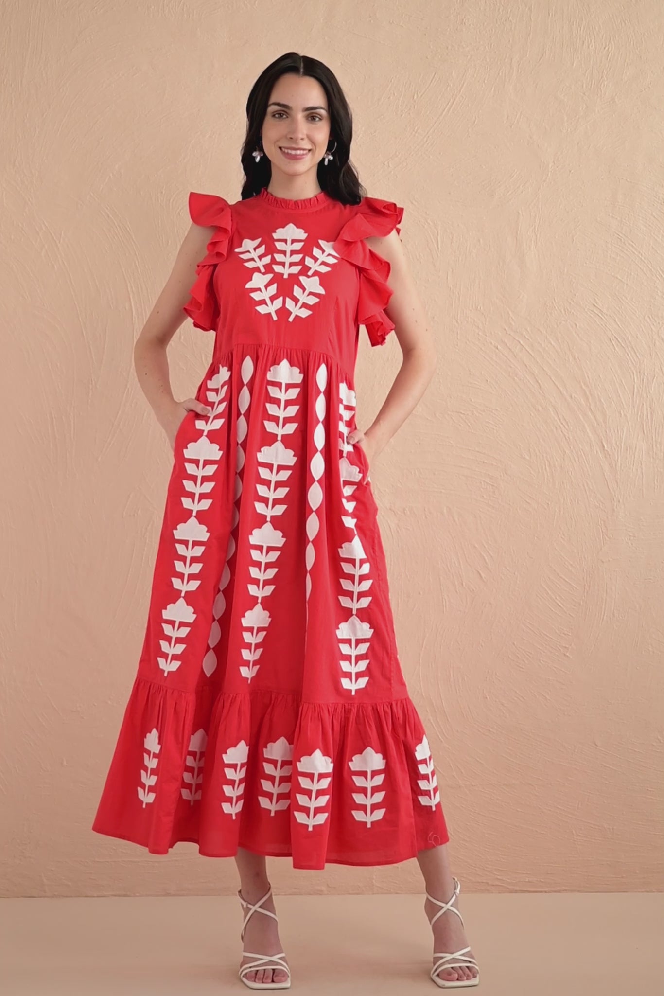 Video of a woman wearing a sleeveless red midi dress with white appliqué embroidery in vertical floral patterns, ruffled shoulders, and a tiered hem.