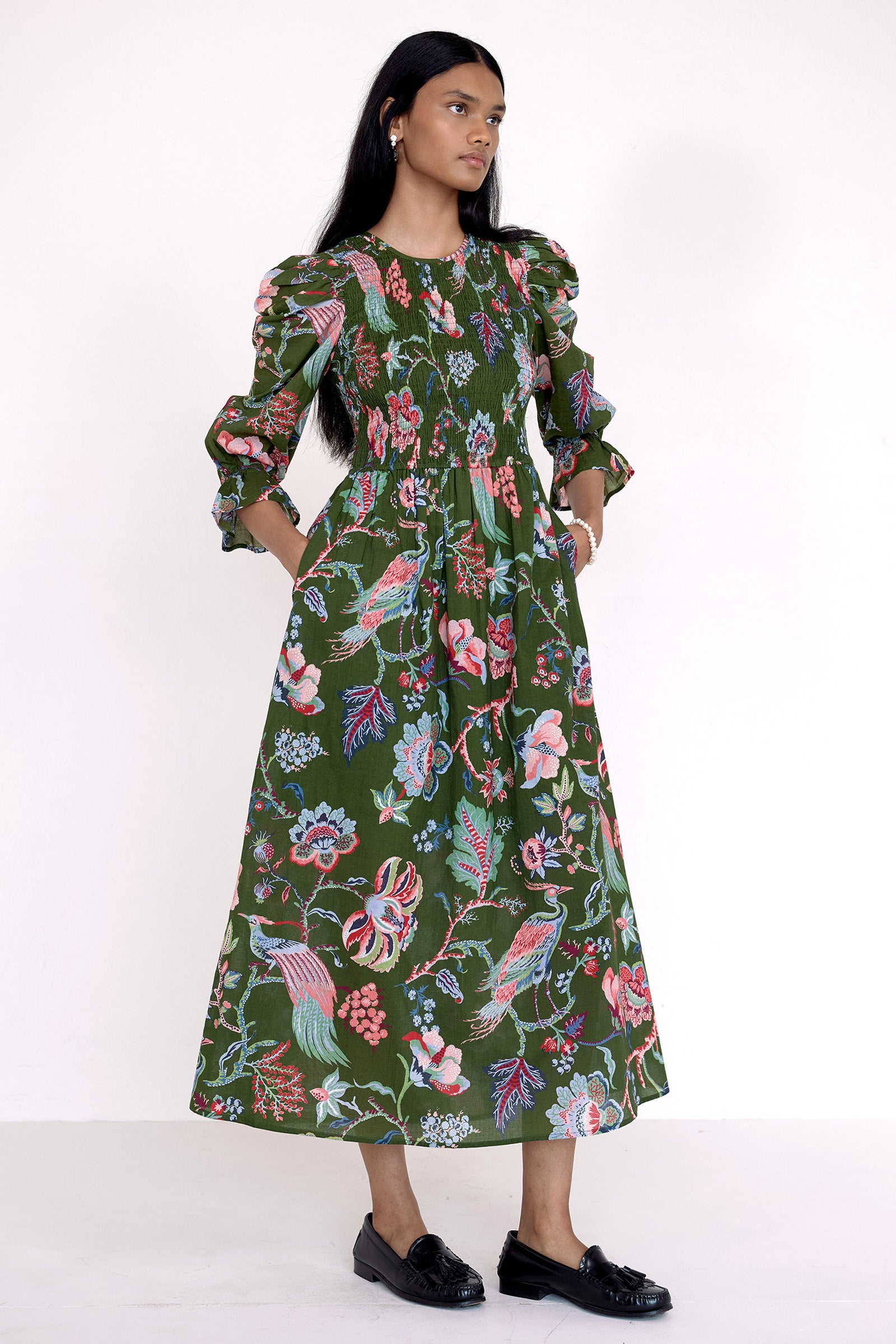 Side view of model standing wearing the Queenie dress in Chintz floral print. It  features a smocked bodice, elbow-length puff sleeves, and a full skirt with pockets.