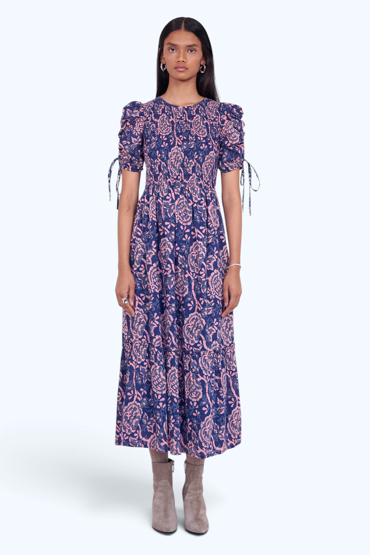 Front view of model standing. She is  wearing the Queenie midi dress in Coromandel print, featuring a smocked bodice, adjustable drawstring sleeves, and a full lined skirt with pockets.