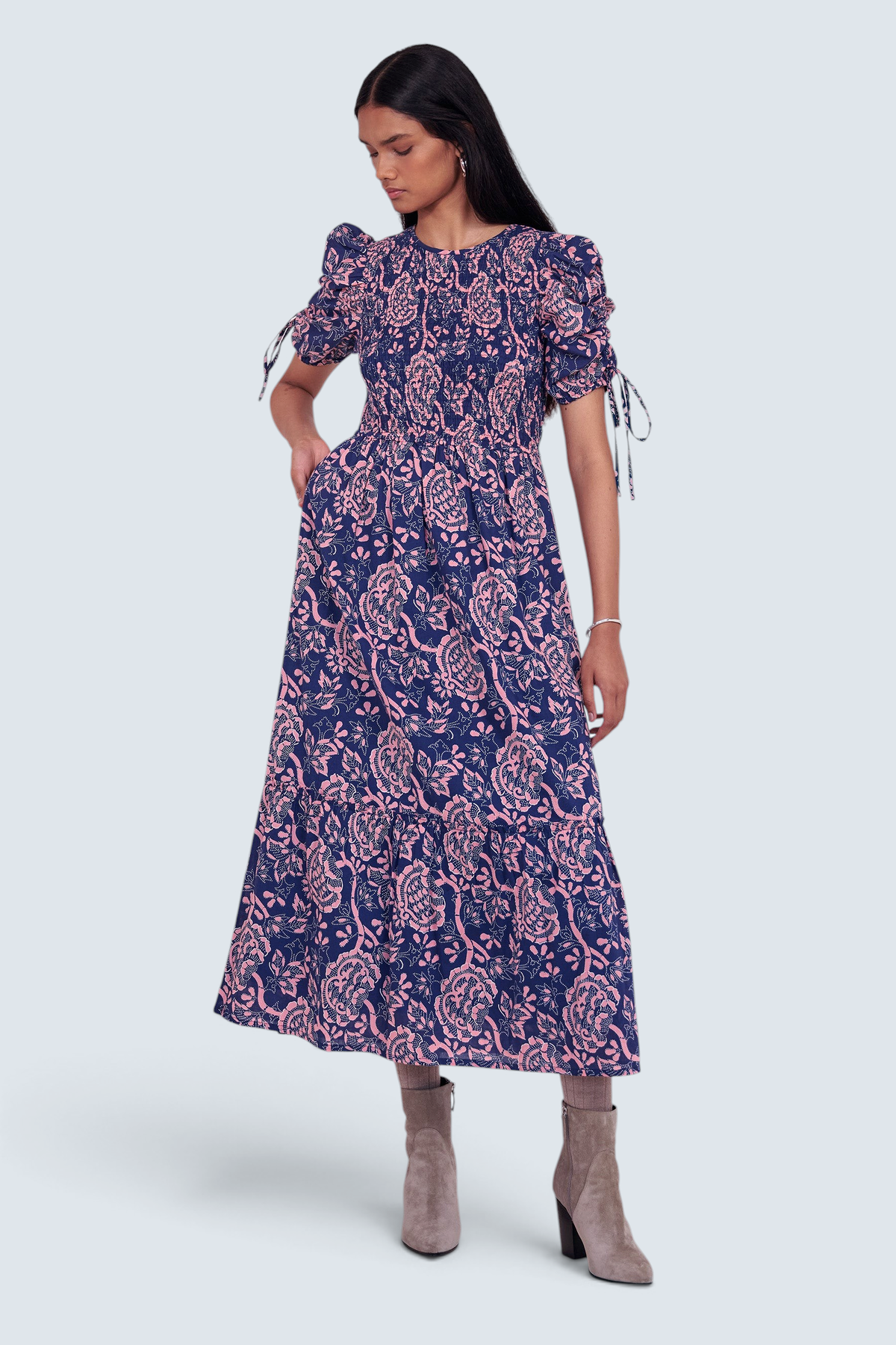 Front view of model standing. She is  wearing the Queenie midi dress in Coromandel print, featuring a smocked bodice, adjustable drawstring sleeves, and a full lined skirt with pockets.