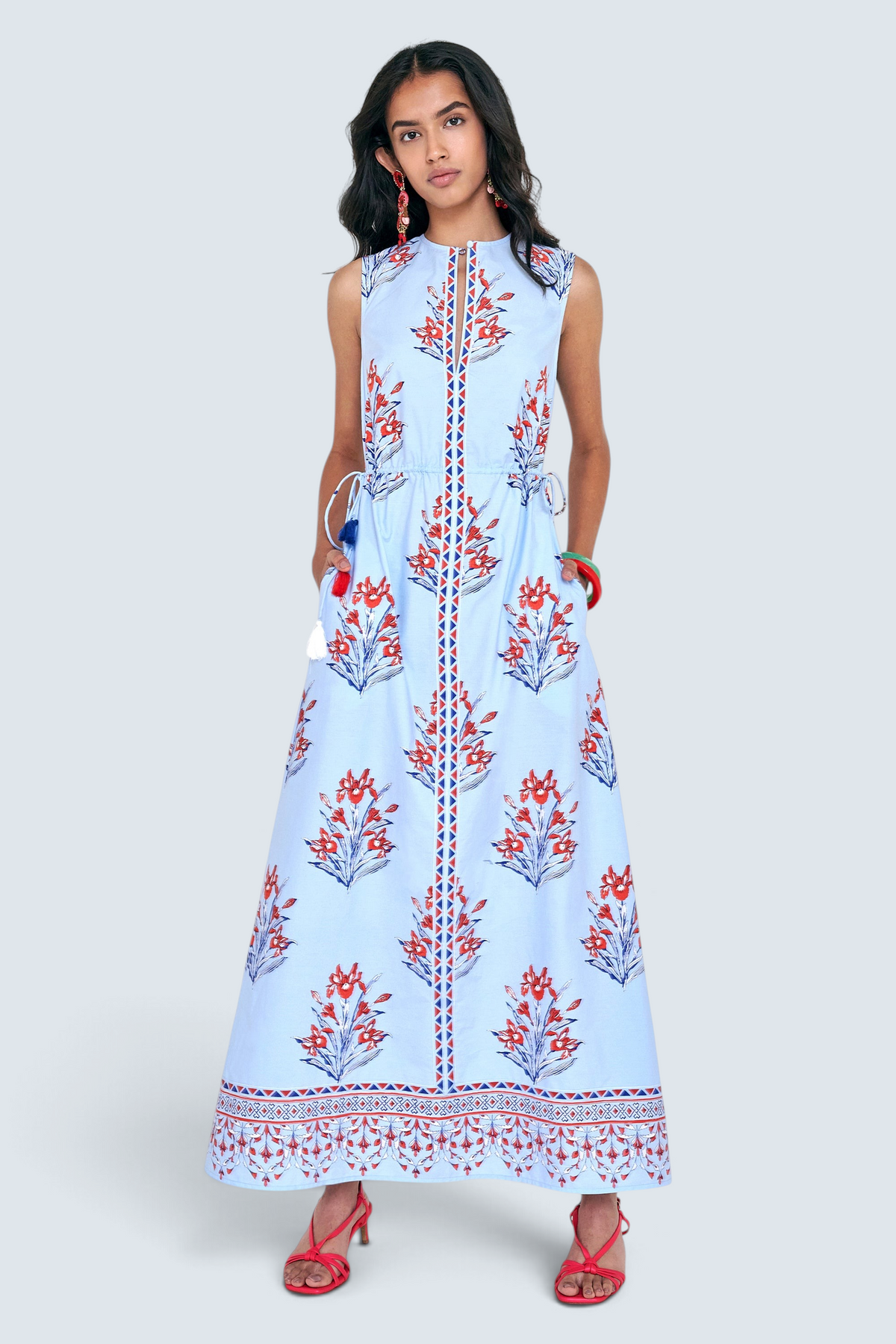 Front view of woman standing. She is  wearing the Rhonda dress in the Brigitte print, featuring a sleek column silhouette, side drawstring details with fringe accents, a keyhole neckline, and iris floral motifs.