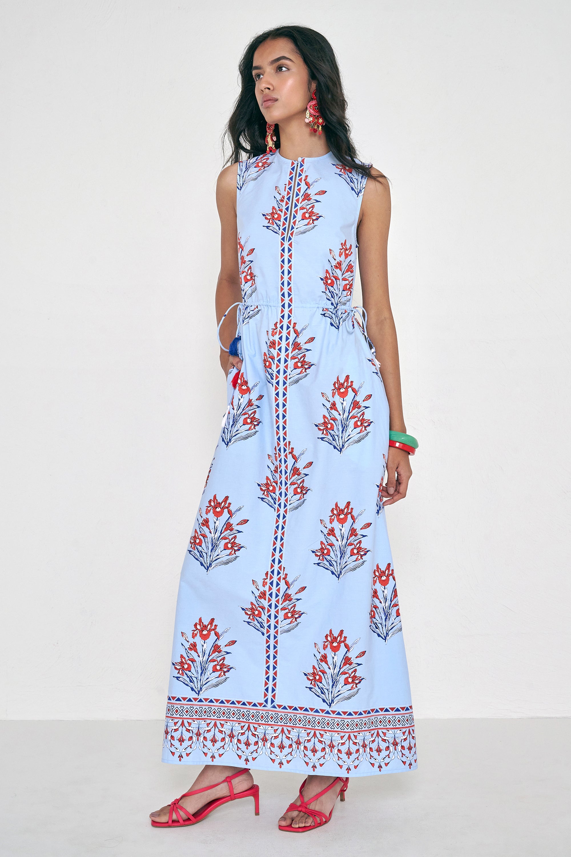 Side view of woman standing. She is  wearing the Rhonda dress in the Brigitte print, featuring a sleek column silhouette, side drawstring details with fringe accents, a keyhole neckline, and iris floral motifs.