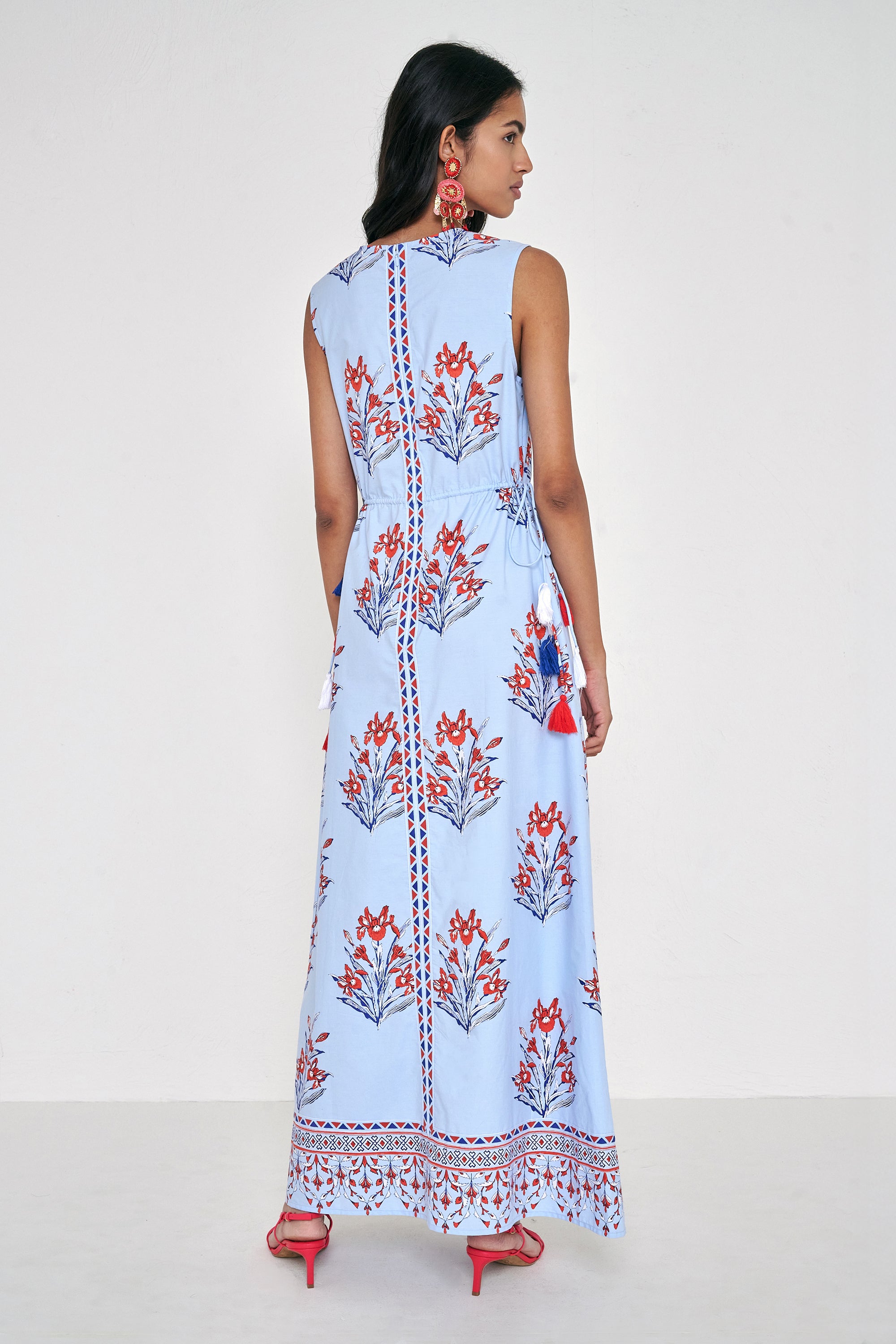 Back view of woman standing. She is  wearing the Rhonda dress in the Brigitte print, featuring a sleek column silhouette, side drawstring details with fringe accents, a keyhole neckline, and iris floral motifs.