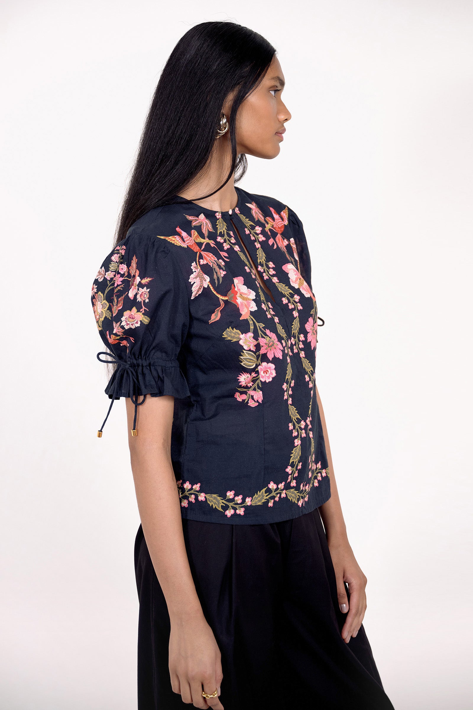 Side view of model standing. She is  wearing the Ricki top in Chintz print, featuring a crewneck with keyhole detail, adjustable drawstring sleeves, and vintage-inspired print placement. Its paired with black pants.