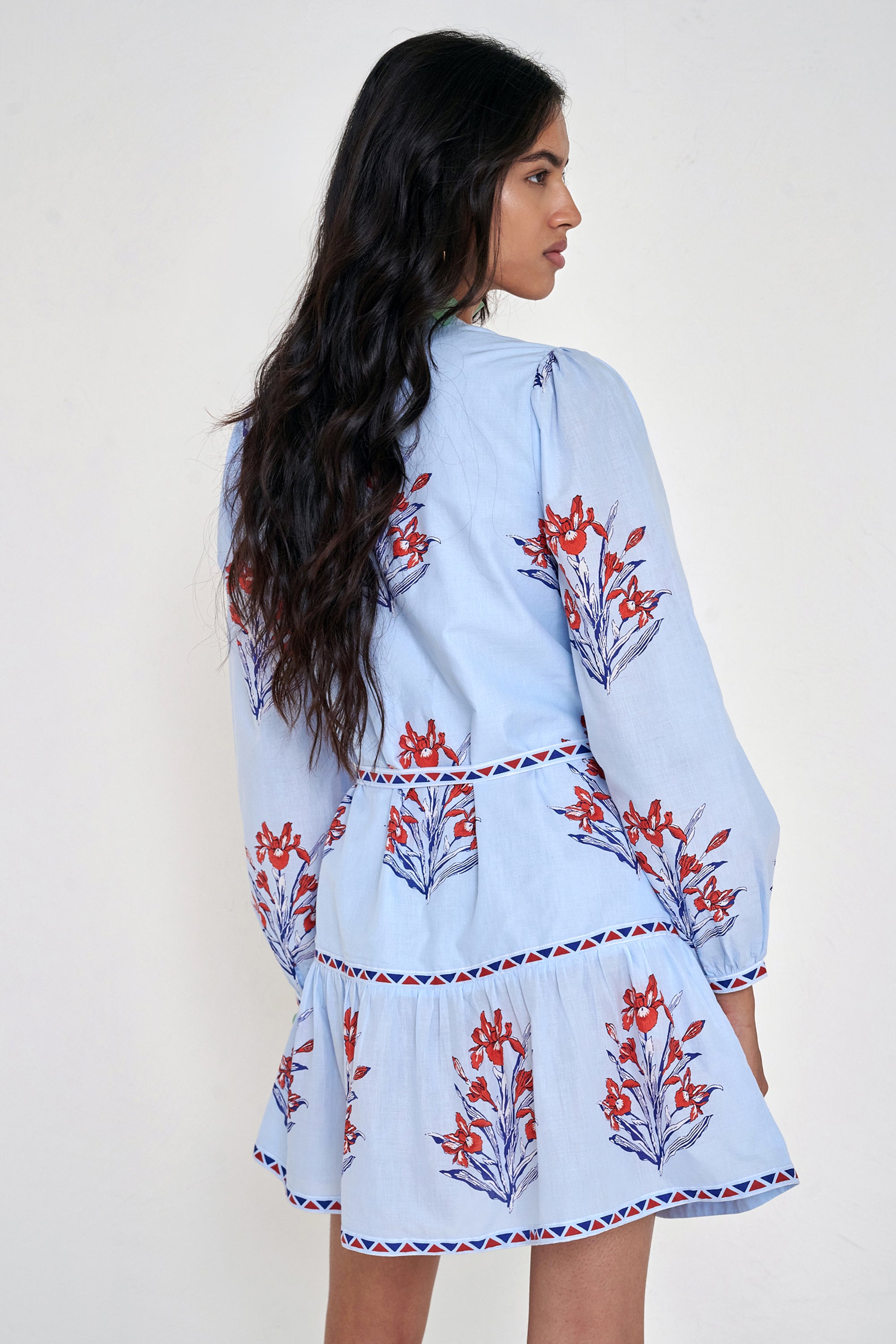 Back view of woman standing. She is wearing the Roma dress in the Brigitte print, featuring a mini shirtdress silhouette, contrast trim details, a detachable belt, and blooming iris floral motifs.