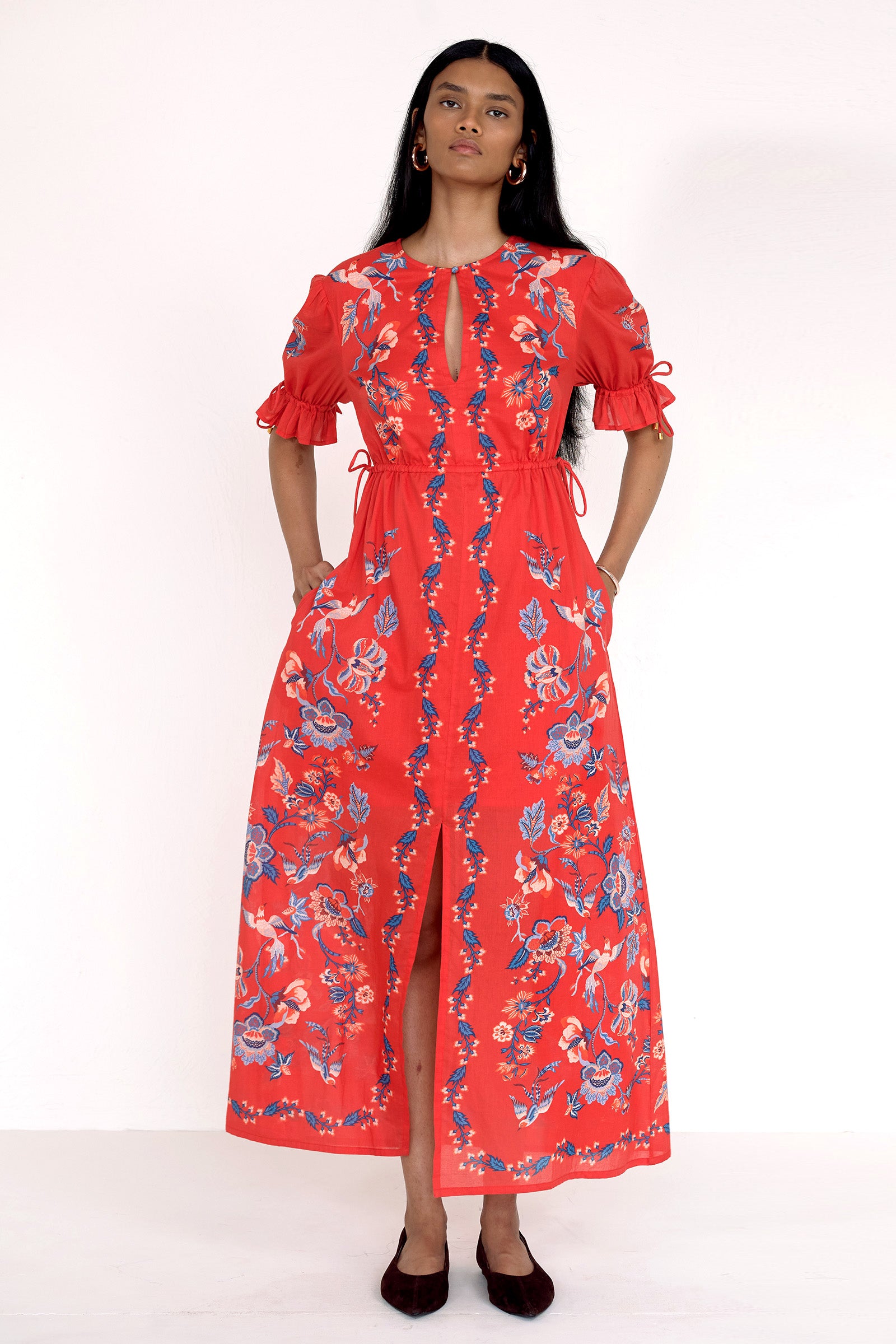 Front view of model standing. She is  wearing the Ruby dress in Chintz print scarlet color with hands in pocket. It features a keyhole neckline, adjustable drawstring waist and sleeves, elbow-length sleeves, and an ankle-length hem.