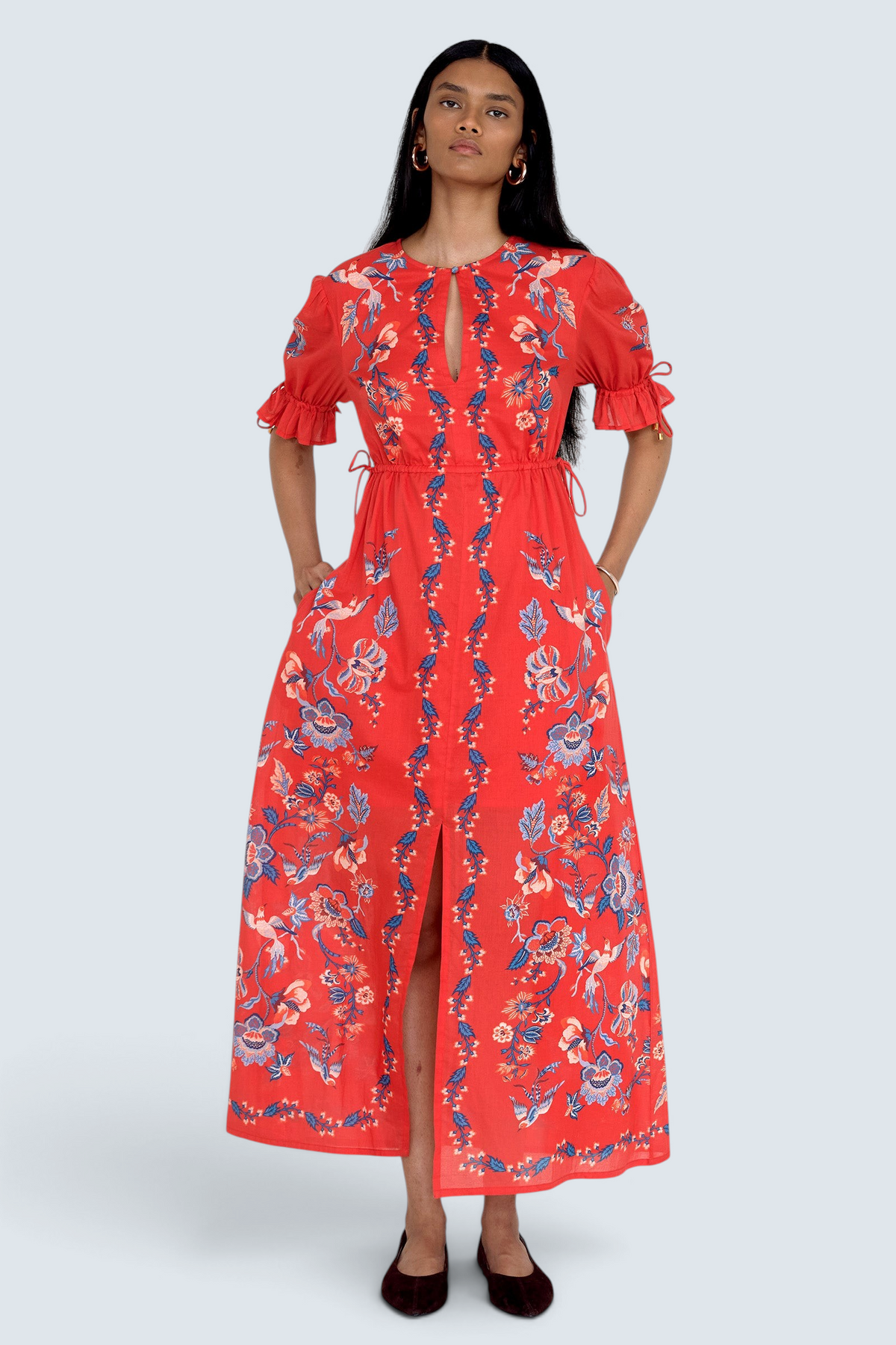 Front view of model standing. She is  wearing the Ruby dress in Chintz print scarlet color with hands in pocket. It features a keyhole neckline, adjustable drawstring waist and sleeves, elbow-length sleeves, and an ankle-length hem.