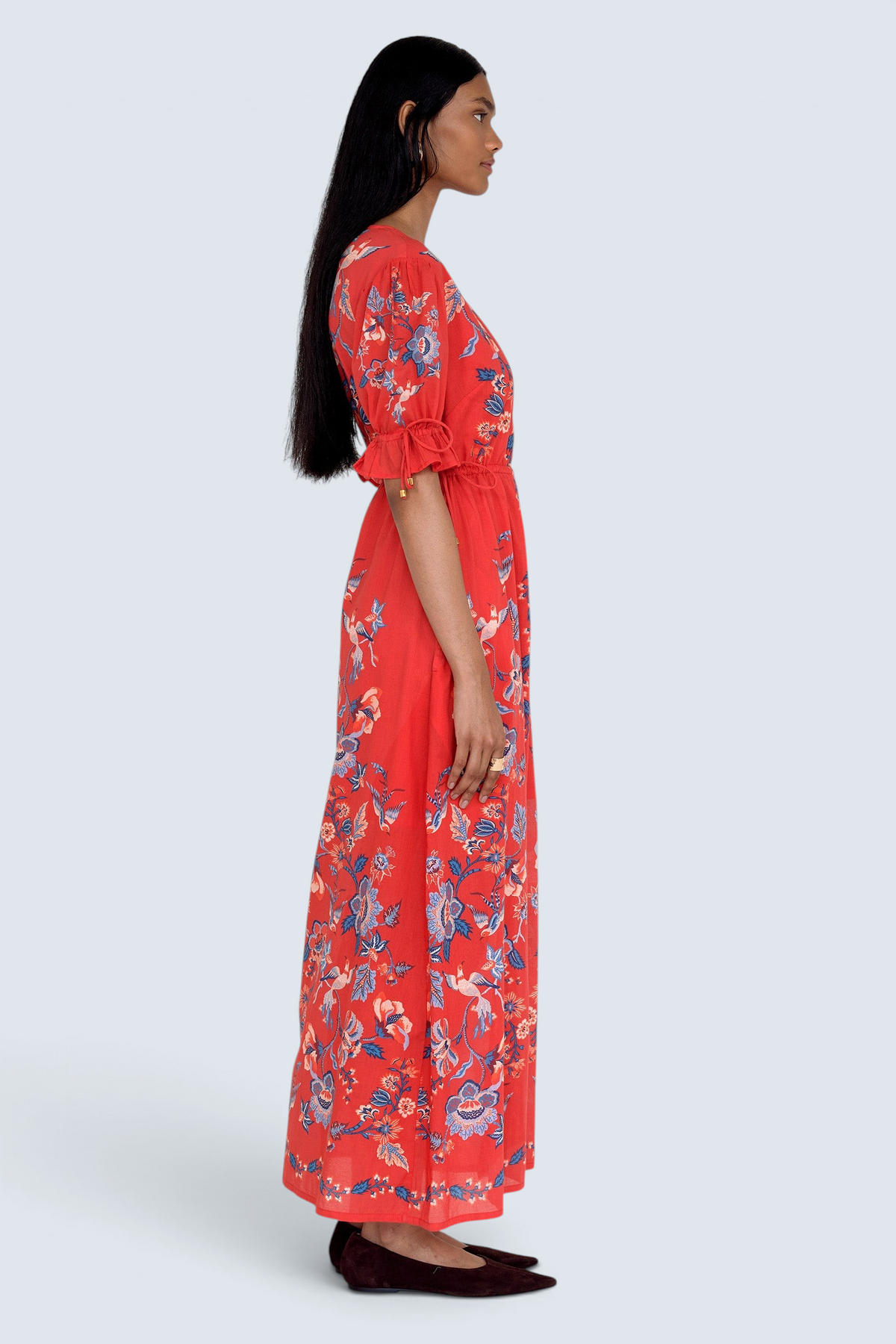 Side view of model standing. She is  wearing the Ruby dress in Chintz print scarlet color. It features a keyhole neckline, adjustable drawstring waist and sleeves, elbow-length sleeves, and an ankle-length hem.