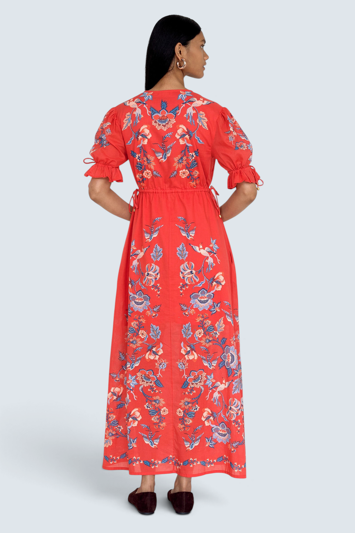 Back view of model standing. She is  wearing the Ruby dress in Chintz print scarlet color. It features a keyhole neckline, adjustable drawstring waist and sleeves, elbow-length sleeves, and an ankle-length hem.