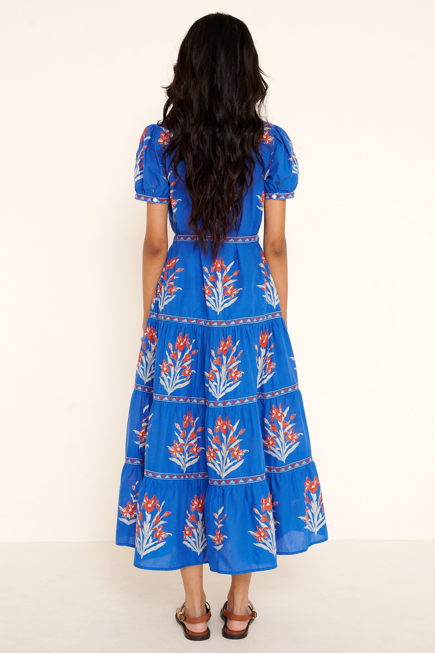 Back view of woman standing. She is  wearing the Saylor dress in the Brigitte print, featuring a full-length shirtdress silhouette, tiered skirt, detachable belt, and hand-screen-printed iris floral motifs.