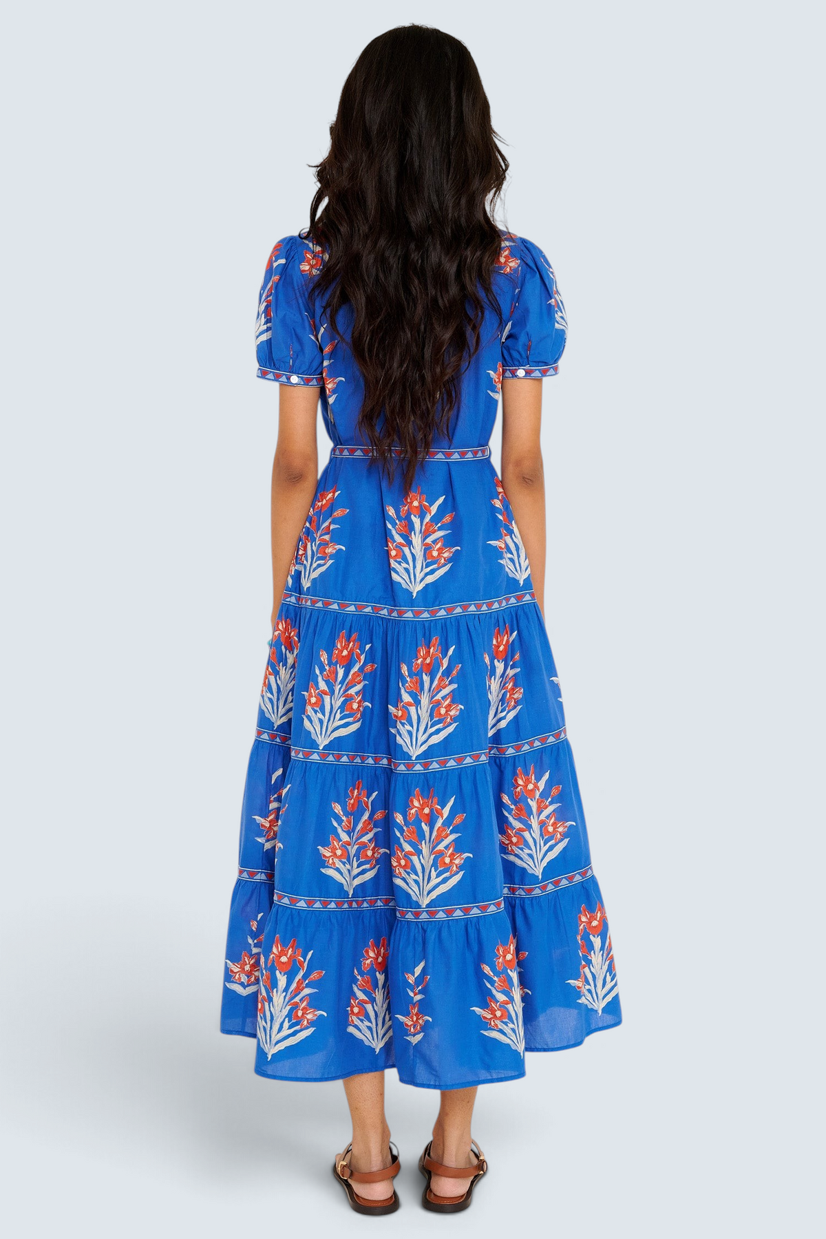 Back view of woman standing. She is  wearing the Saylor dress in the Brigitte print, featuring a full-length shirtdress silhouette, tiered skirt, detachable belt, and hand-screen-printed iris floral motifs.