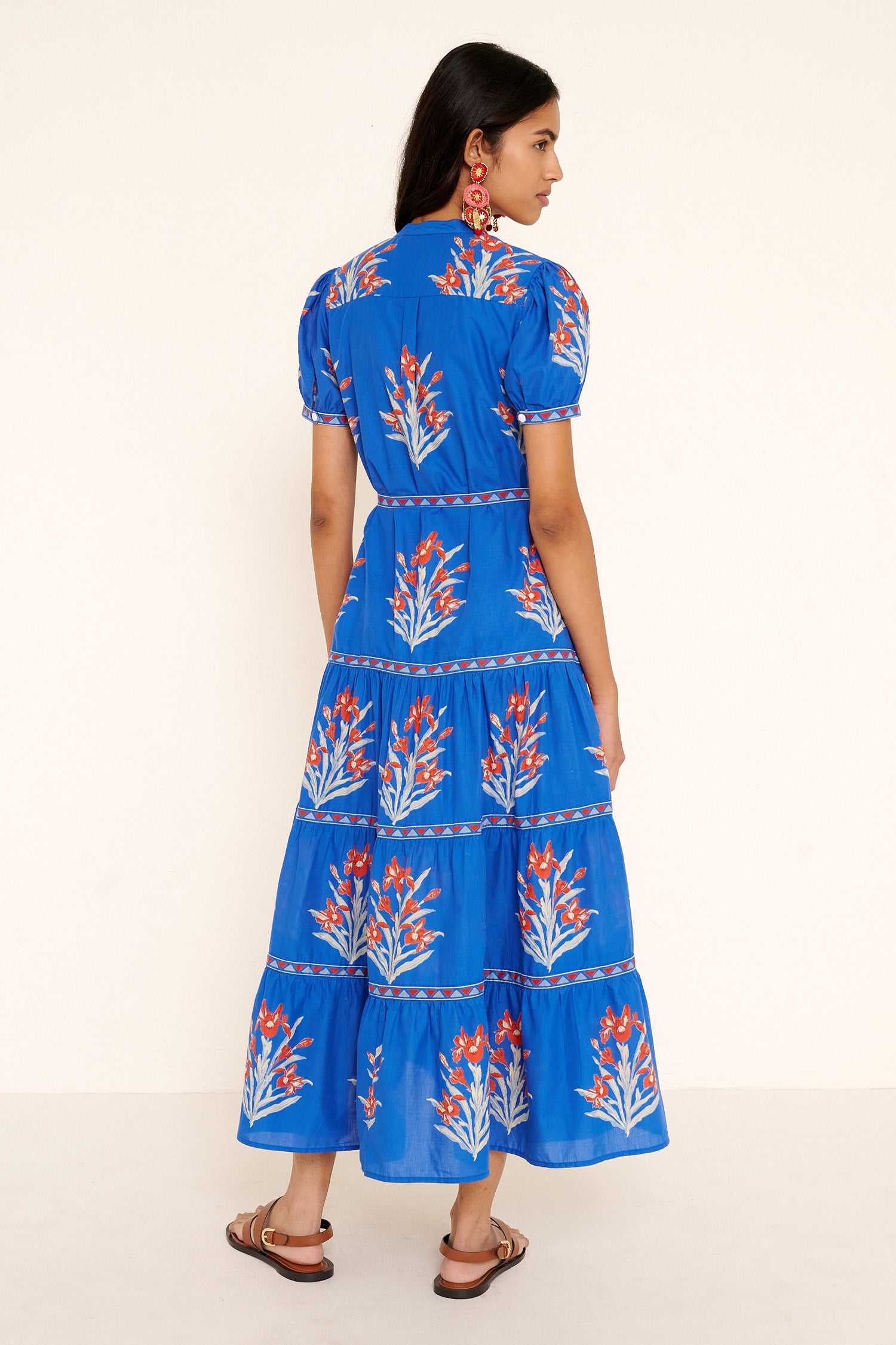 Back view of woman standing. She is  wearing the Saylor dress in the Brigitte print, featuring a full-length shirtdress silhouette, tiered skirt, detachable belt, and hand-screen-printed iris floral motifs.