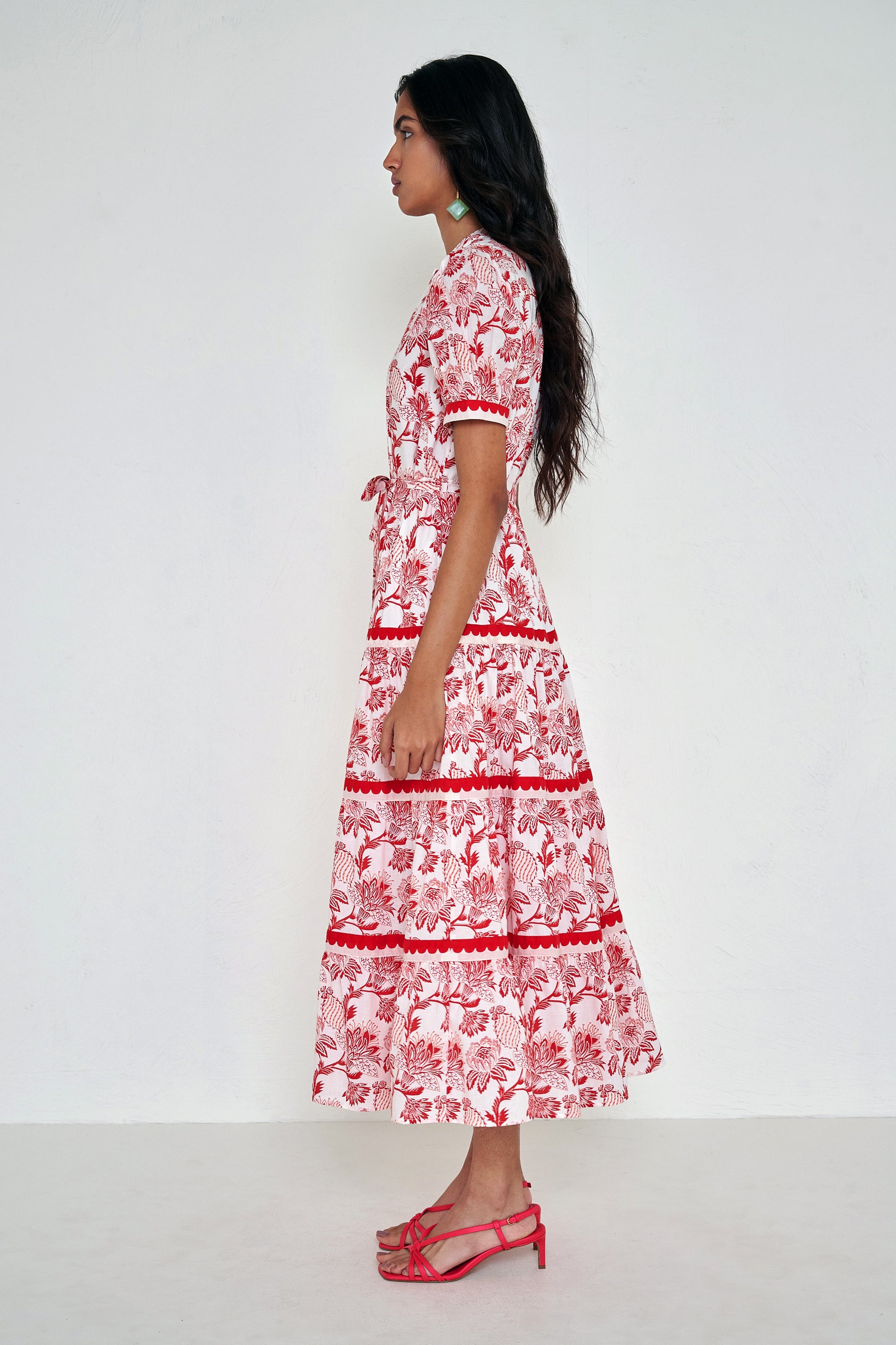 Side view of woman standing. She is  wearing the Saylor dress in the Lizzie floral print, featuring a full-length shirtdress silhouette, tiered skirt, detachable belt, and winding floral vine motifs.