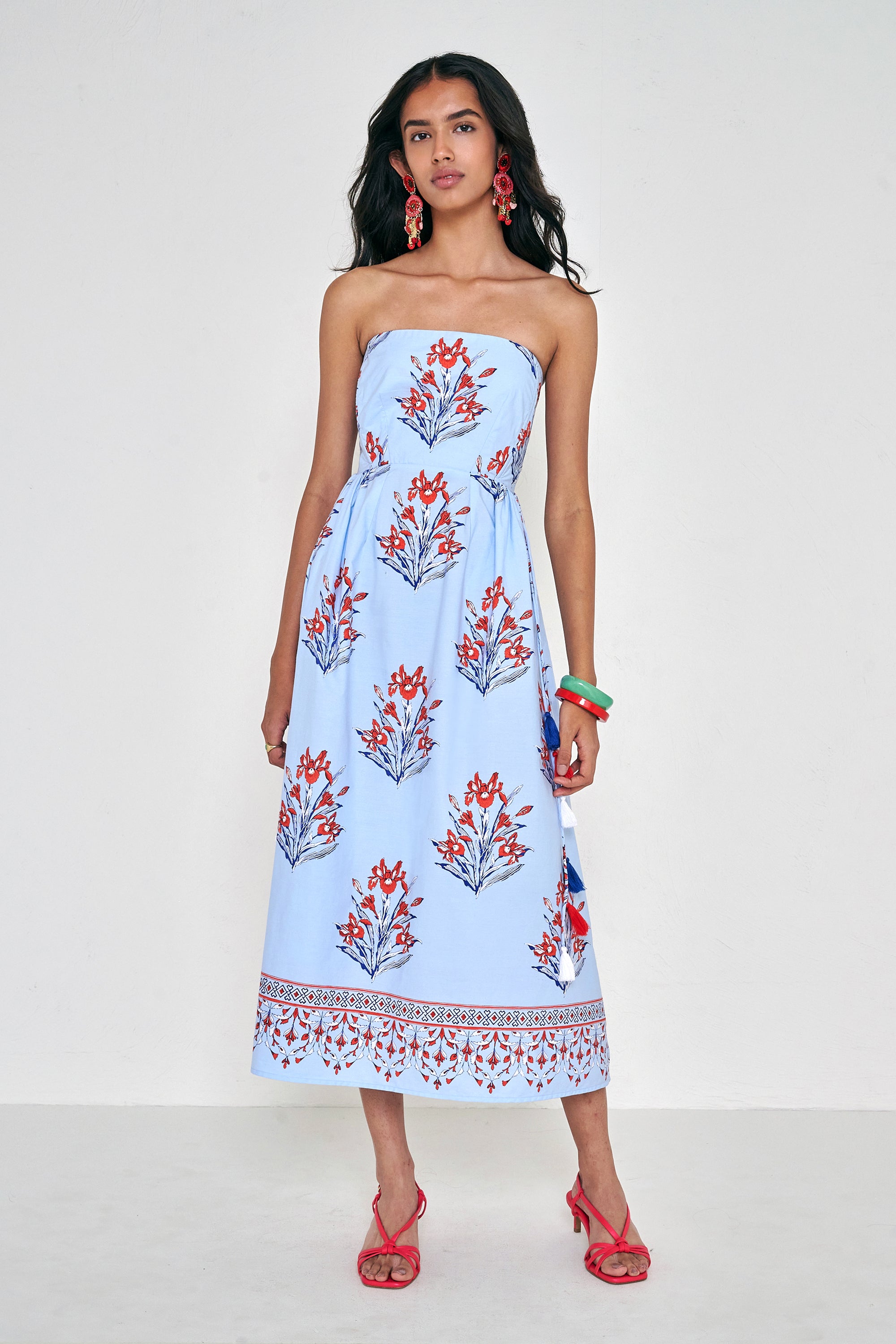 Front view of woman standing. She is  wearing the Simona dress in the Brigitte floral print, featuring a strapless bodice, full gathered skirt, and red, white, and blue iris motifs.