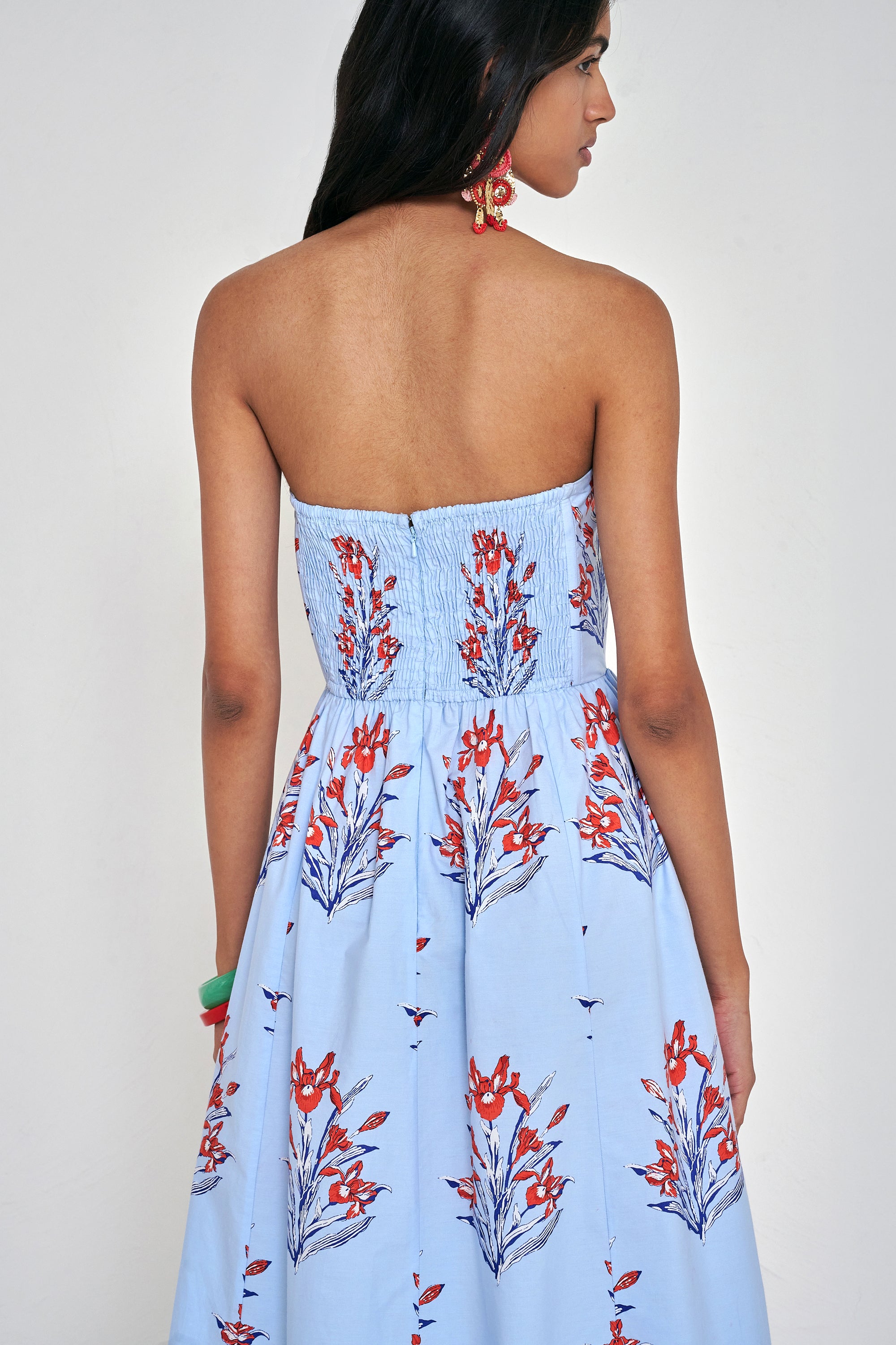 Back view close up of woman standing. She is  wearing the Simona dress in the Brigitte floral print, featuring a strapless bodice, full gathered skirt, and red, white, and blue iris motifs.