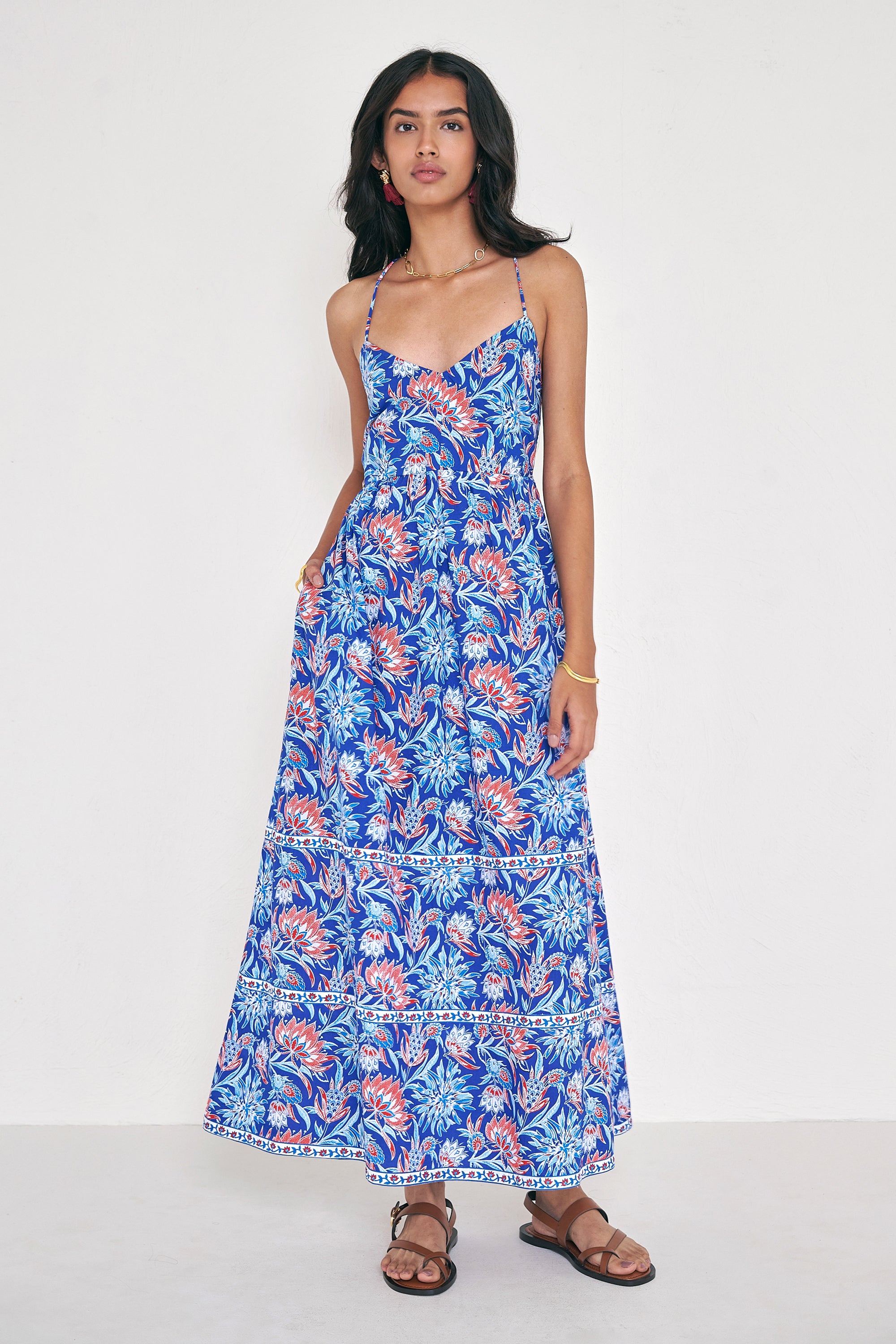 Front view of woman standing. She is  wearing the Sofia dress in the Philippa print, featuring a strappy sundress silhouette, gathered ankle-length skirt, and red, white, and blue wildflower motifs inspired by Provence.