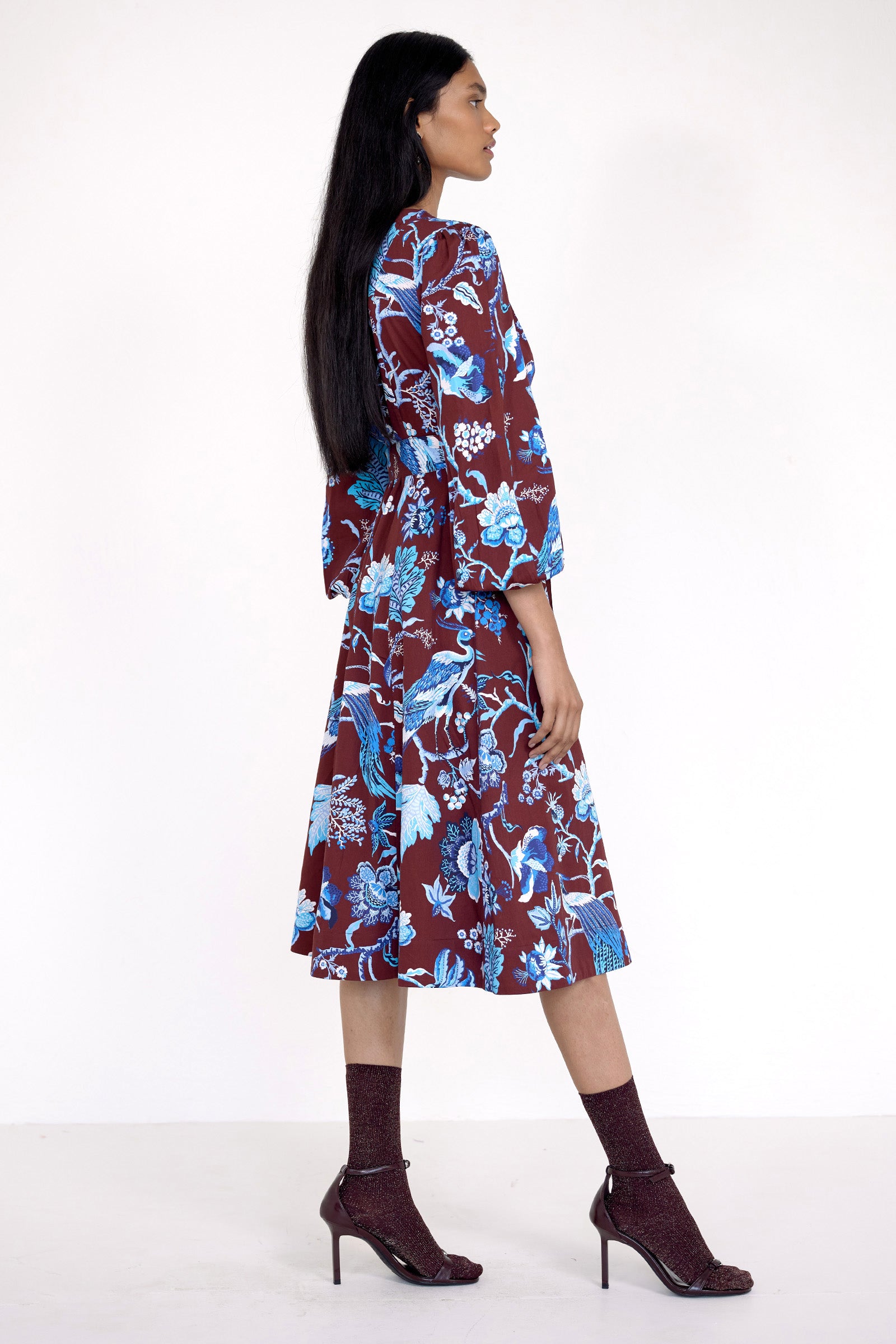 Side view of model standing. She is  wearing the Zora dress in Chintz print, featuring a V-neck, lantern sleeves, princess seams, a gathered fit-and-flare skirt, fabric belt, and side-seam pockets.