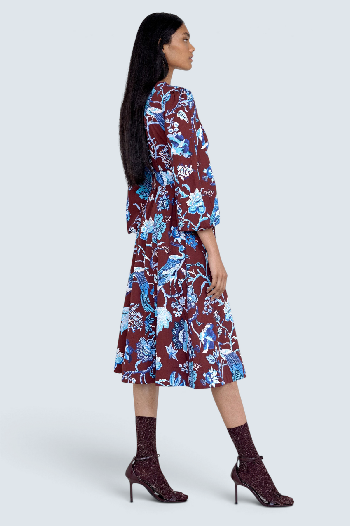 Side view of model standing. She is  wearing the Zora dress in Chintz print, featuring a V-neck, lantern sleeves, princess seams, a gathered fit-and-flare skirt, fabric belt, and side-seam pockets.
