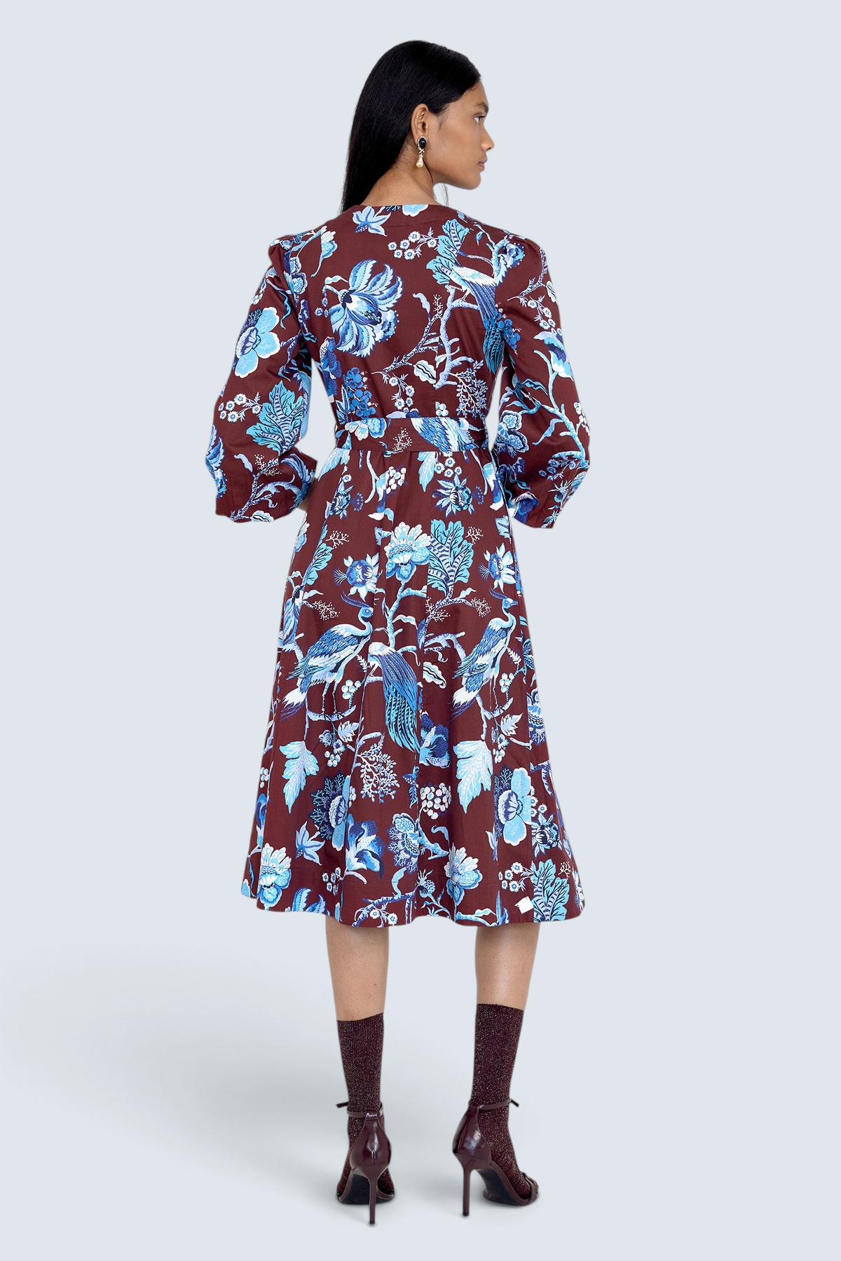 Back view of model standing. She is  wearing the Zora dress in Chintz print, featuring a V-neck, lantern sleeves, princess seams, a gathered fit-and-flare skirt, fabric belt, and side-seam pockets.